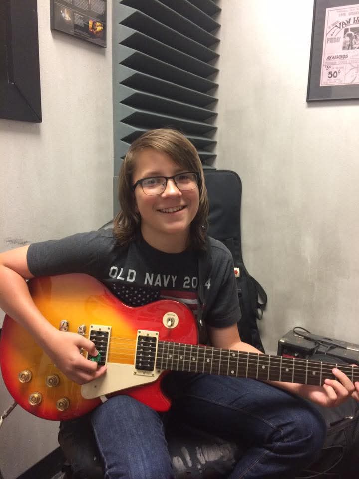 Person with glasses smiling, holding a red and yellow electric guitar, sitting in a room with soundproof panels.