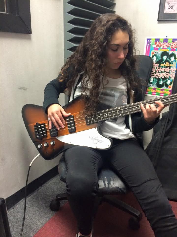 Young woman with curly hair playing an electric bass guitar in a room with soundproofing panels.