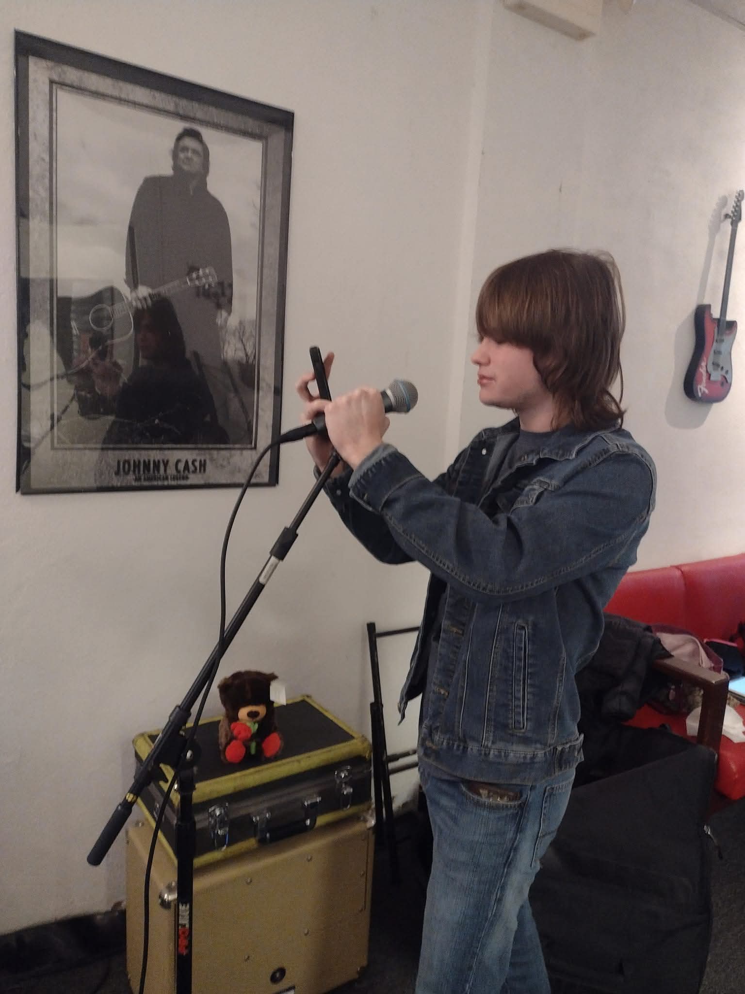 Young person with brown hair in a denim jacket and jeans holding a microphone, standing near a guitar on the wall.