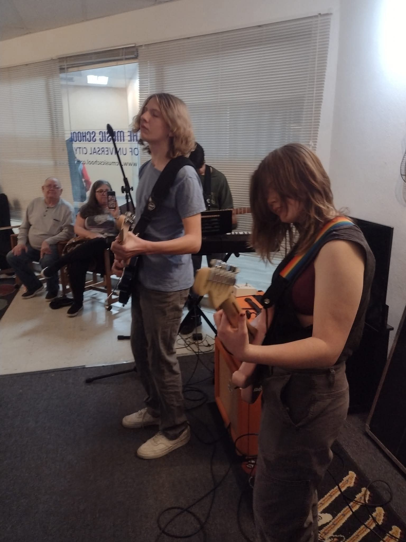 Two women playing guitars in a room with seated audience members and musical equipment.