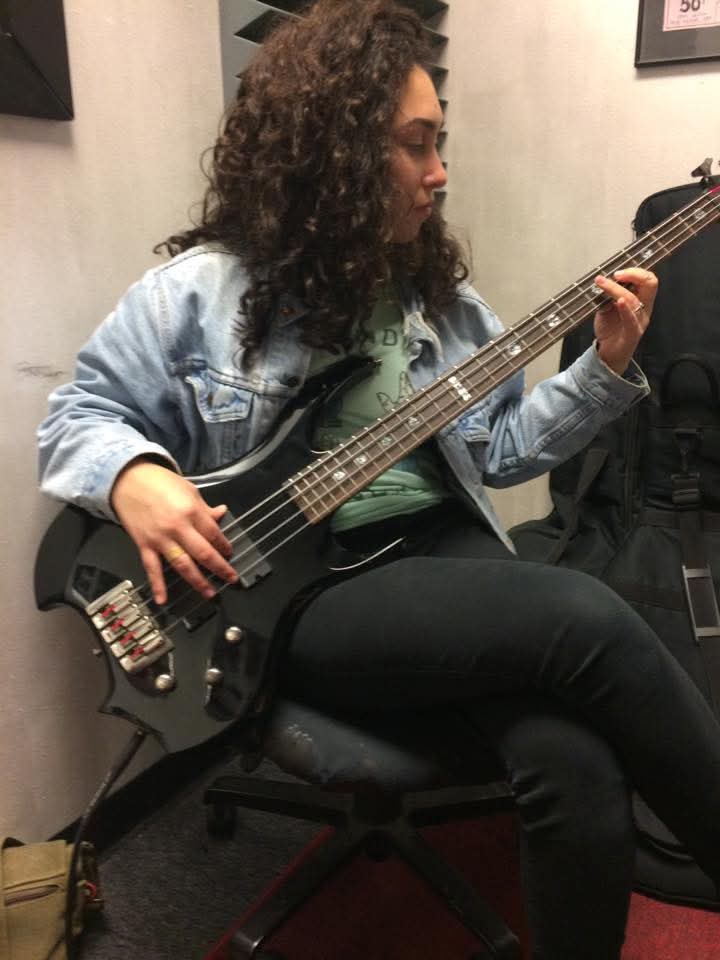 Woman with curly hair playing a black bass guitar while seated, wearing a denim jacket and black pants.