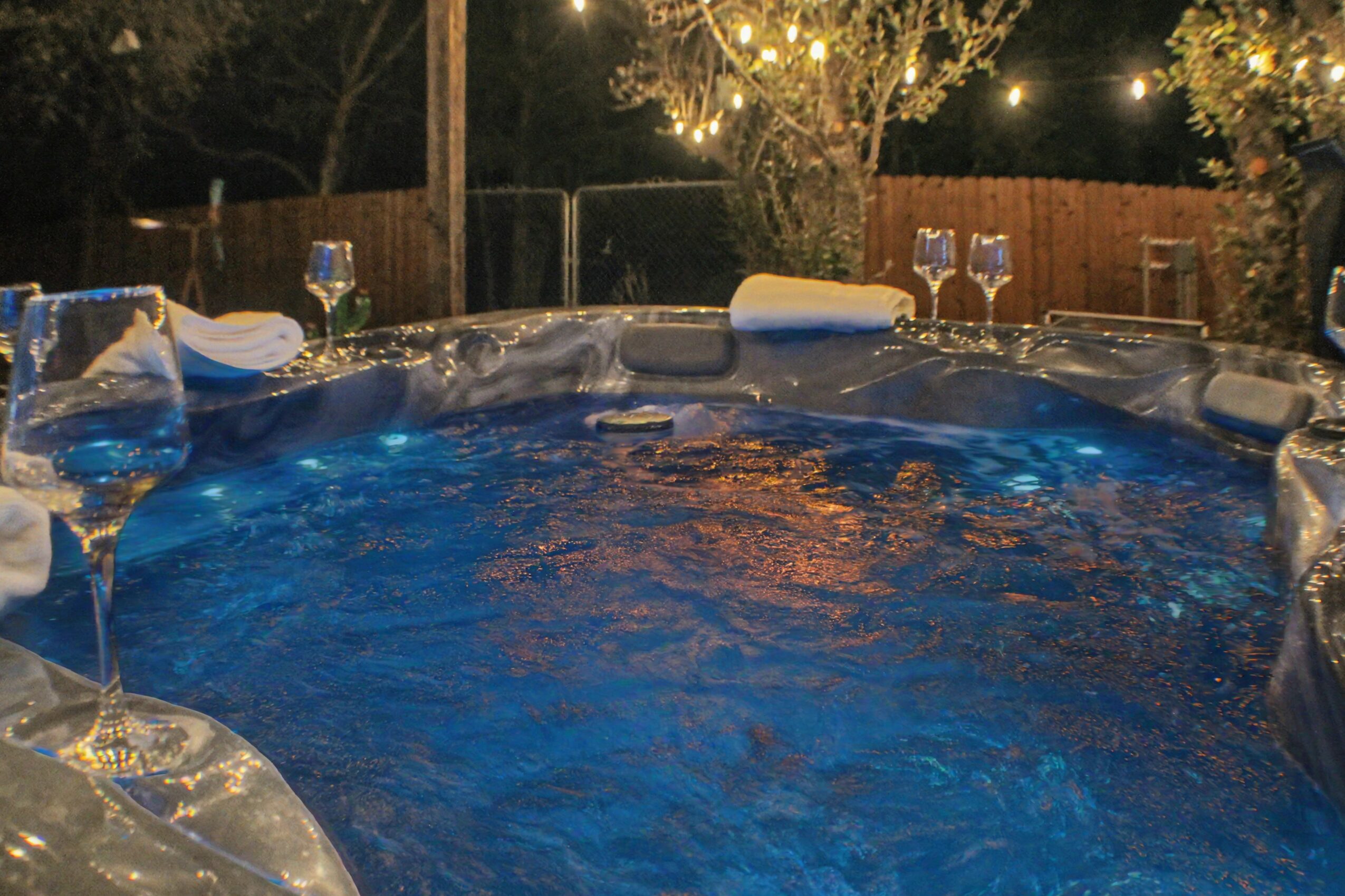 Hot tub filled with water, surrounded by glasses and tableware, outdoor setting with string lights and trees at night.