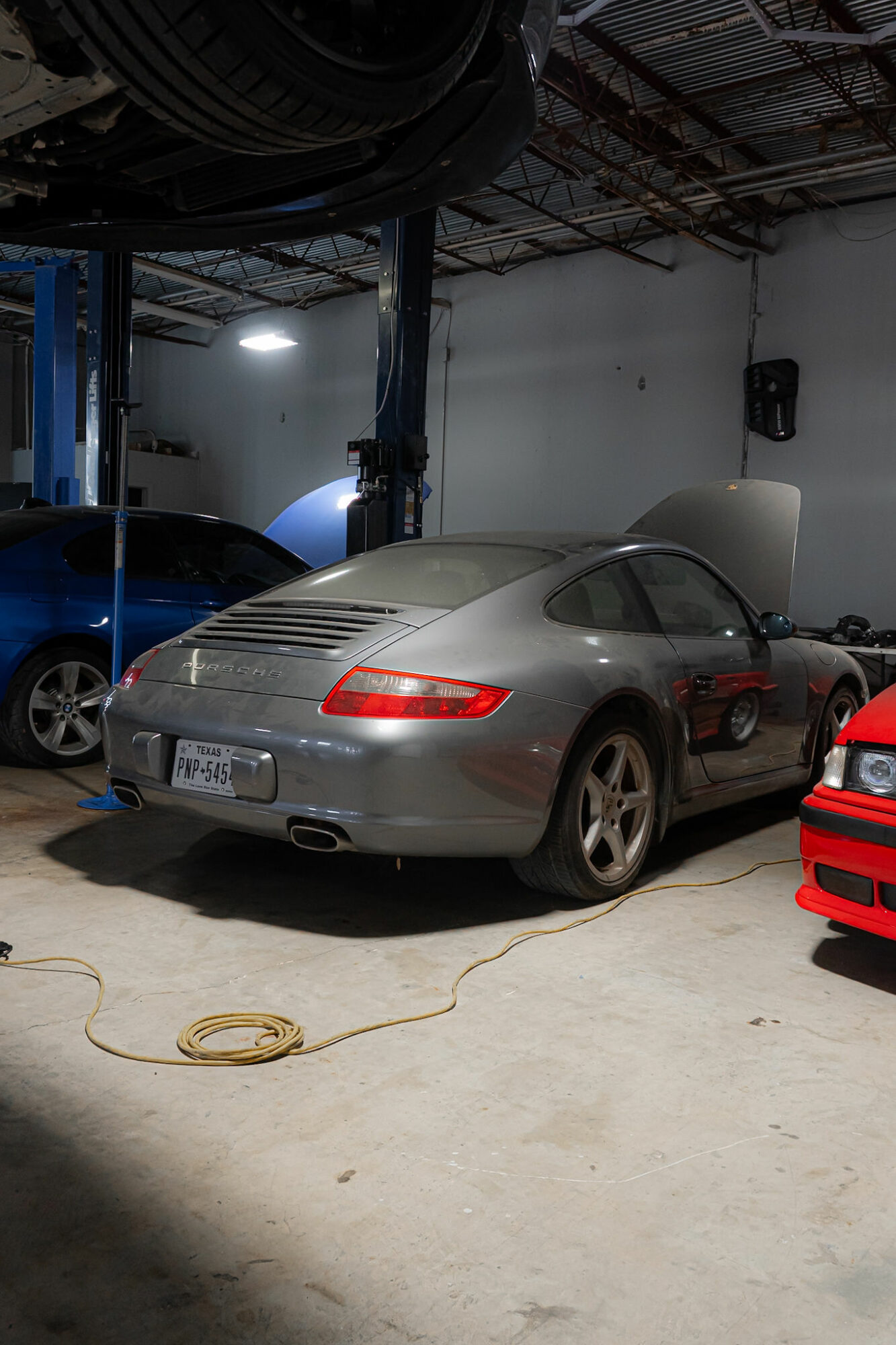Silver sports car with open hood parked indoors, surrounded by other vehicles and equipment, with a yellow extension cord on the floor.