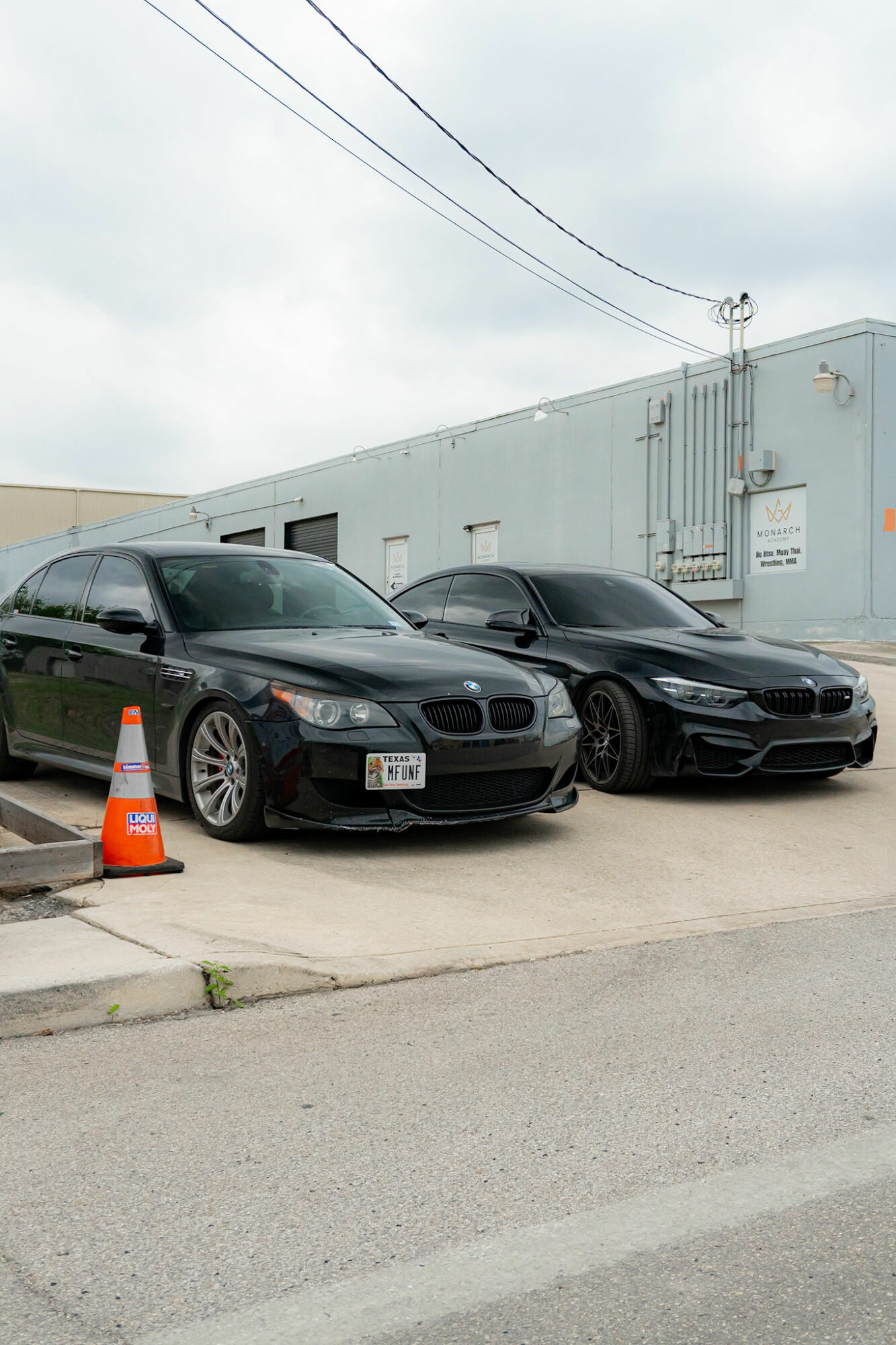 Two black cars parked side by side in front of a light blue industrial building, with an orange traffic cone nearby.