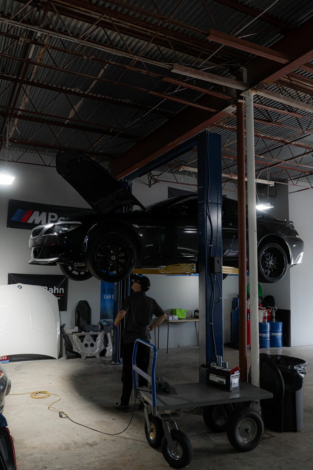 Black sports car on a lift inside a garage with a mechanic working below, equipment and tools around.