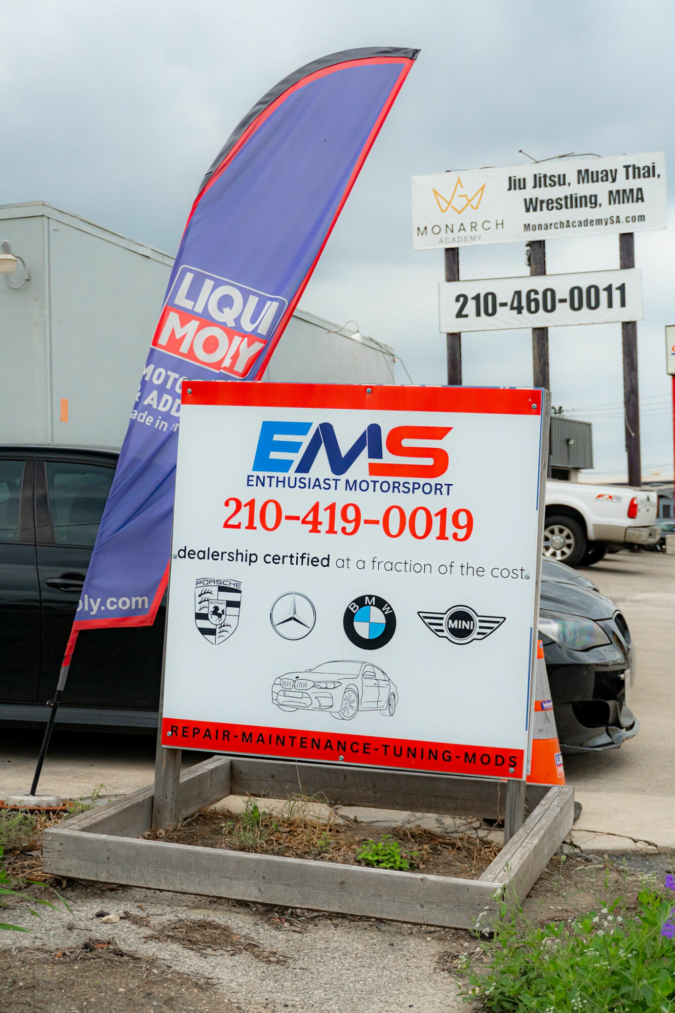 Signboard for Enthusiast Motorsport with logos of car brands, contact number, and red banner at bottom, outdoors.