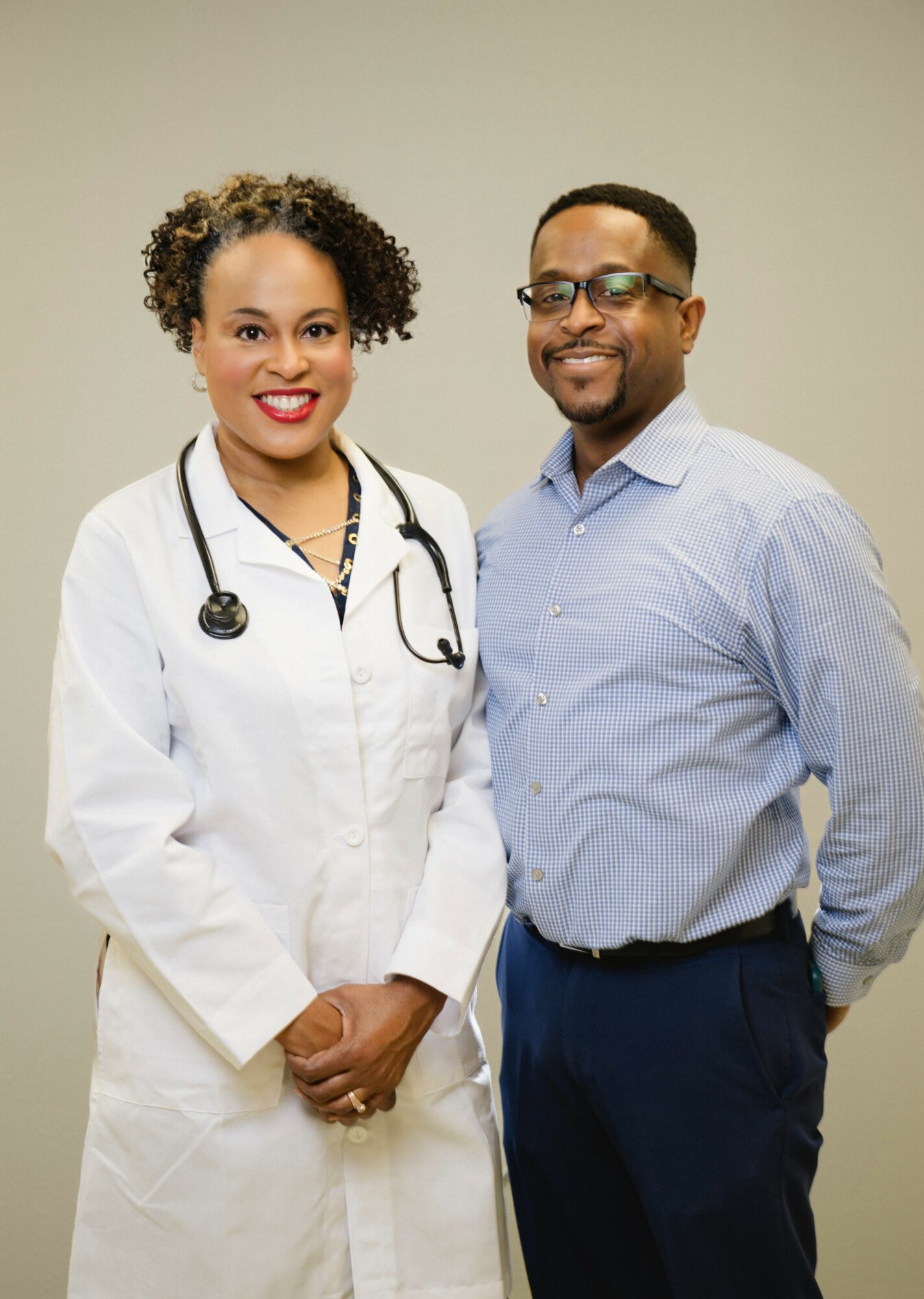 Female doctor in white coat with stethoscope, smiling, standing next to male colleague in light blue shirt, smiling, in neutral background.