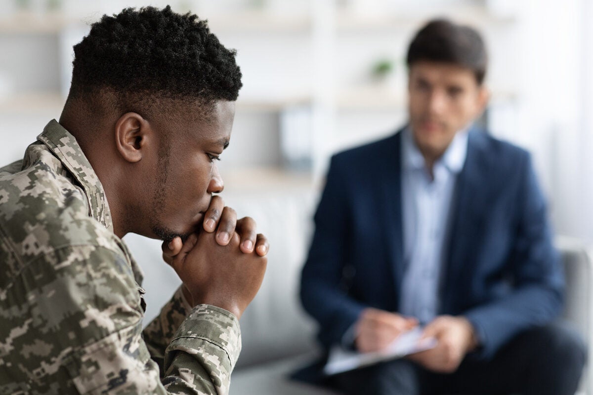 Young man in camouflage shirt with hands clasped, talking to a person in a suit in a bright office.