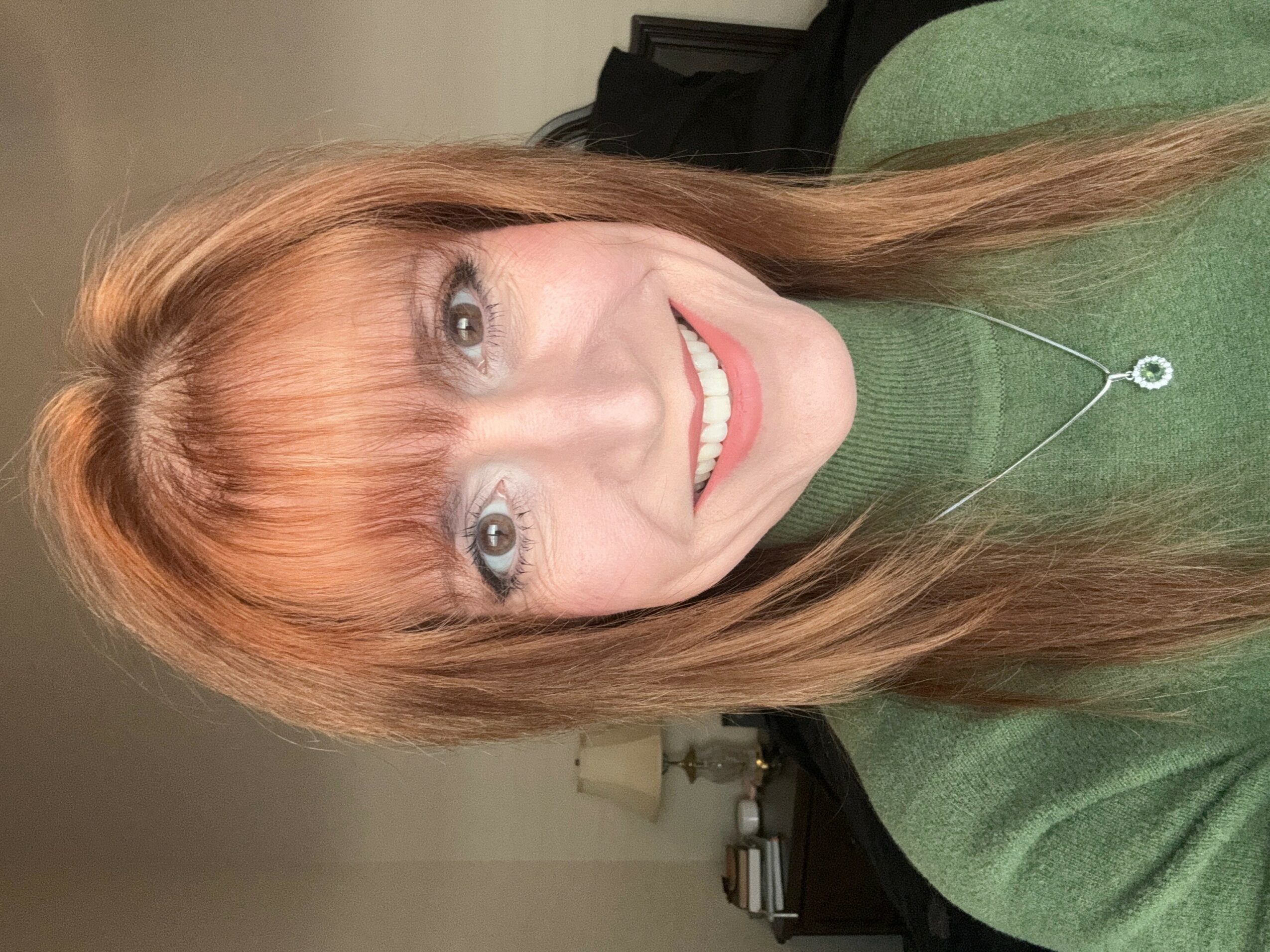 Woman with long reddish hair, smiling, wearing a green top and a necklace, indoors with a lamp and furniture in background.