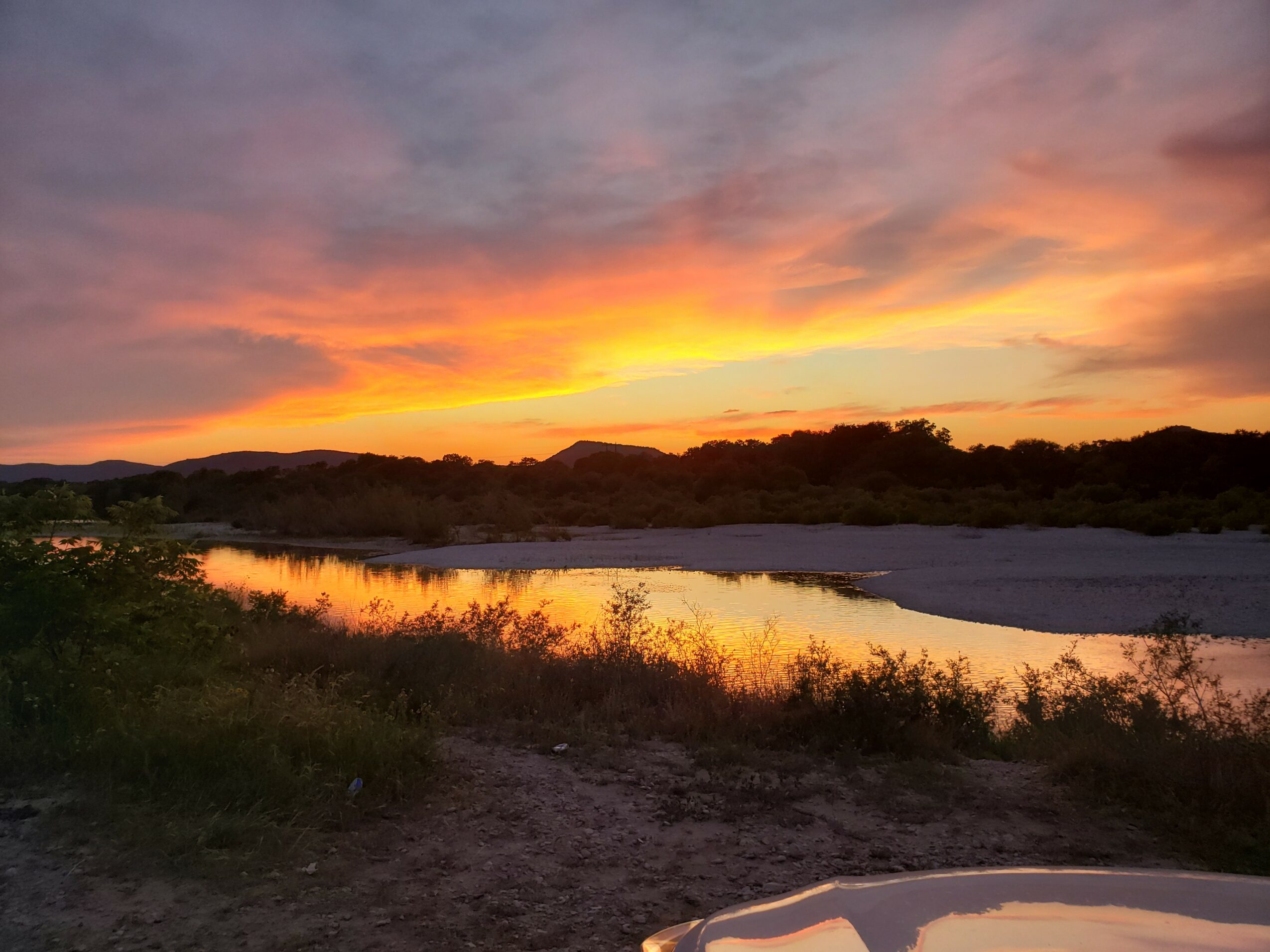 Sunset over a river with colorful sky and silhouetted trees, reflecting on the water, with a sandy foreground.