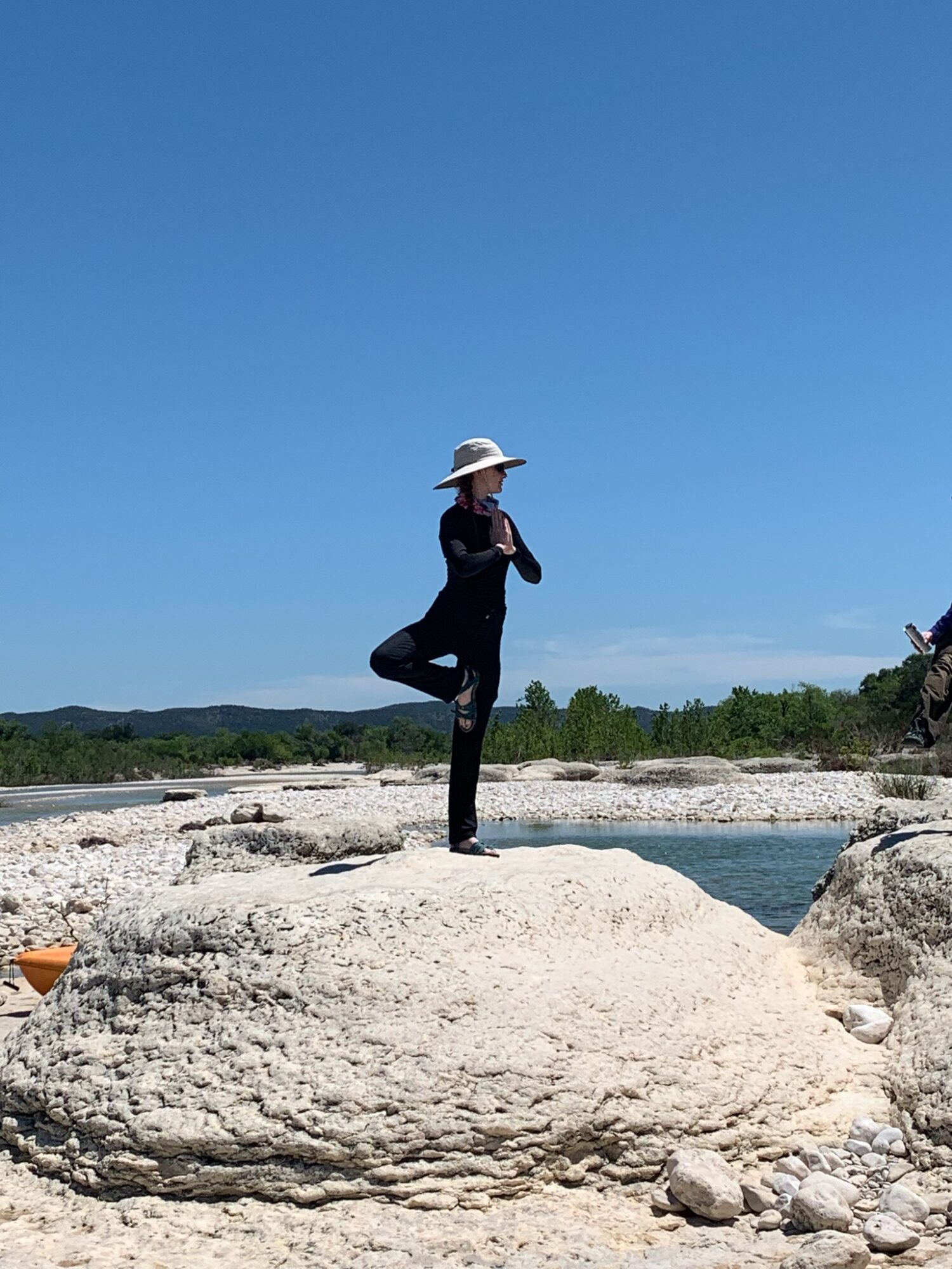 Person standing on a large rock near water, wearing a wide-brimmed hat and dark clothing, with a clear blue sky.