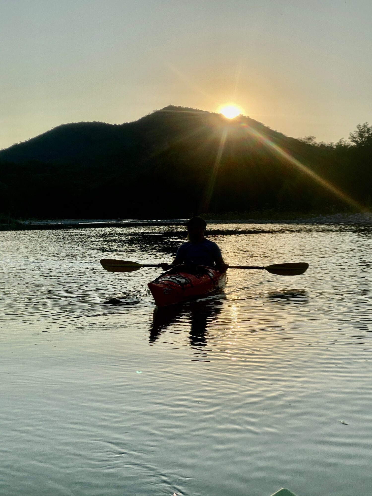 Person kayaking on a calm body of water during sunset with a mountain in the background.