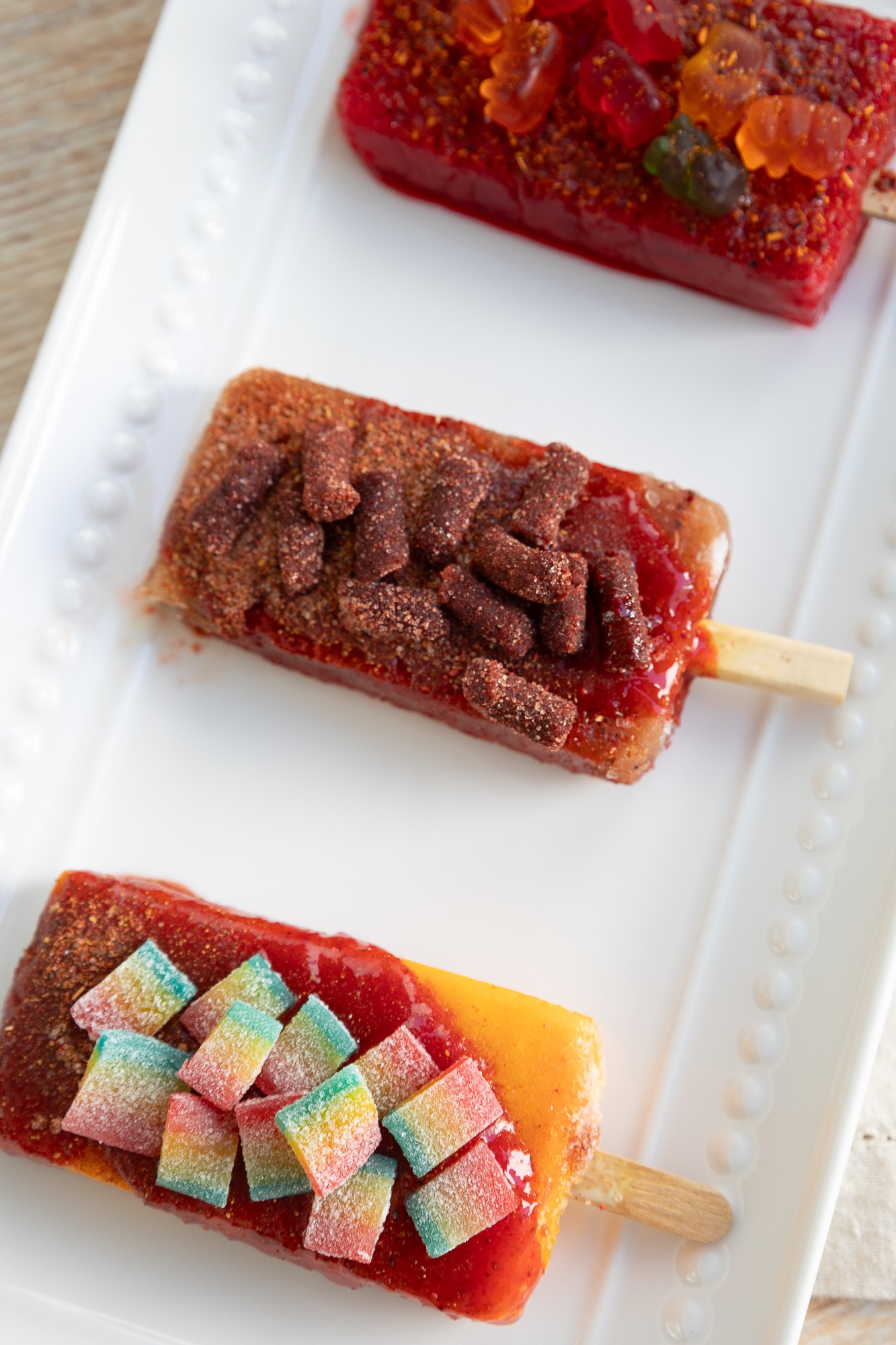 Three colorful fruit popsicles on a white rectangular plate, each with different toppings and flavors.