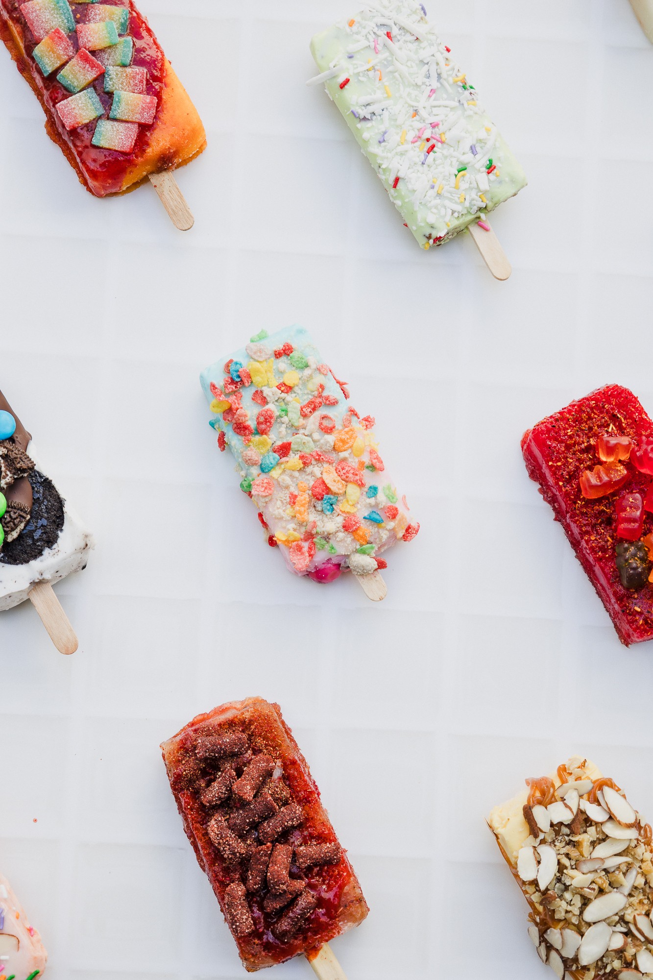 Six colorful ice cream bars with various toppings on a white tiled surface.