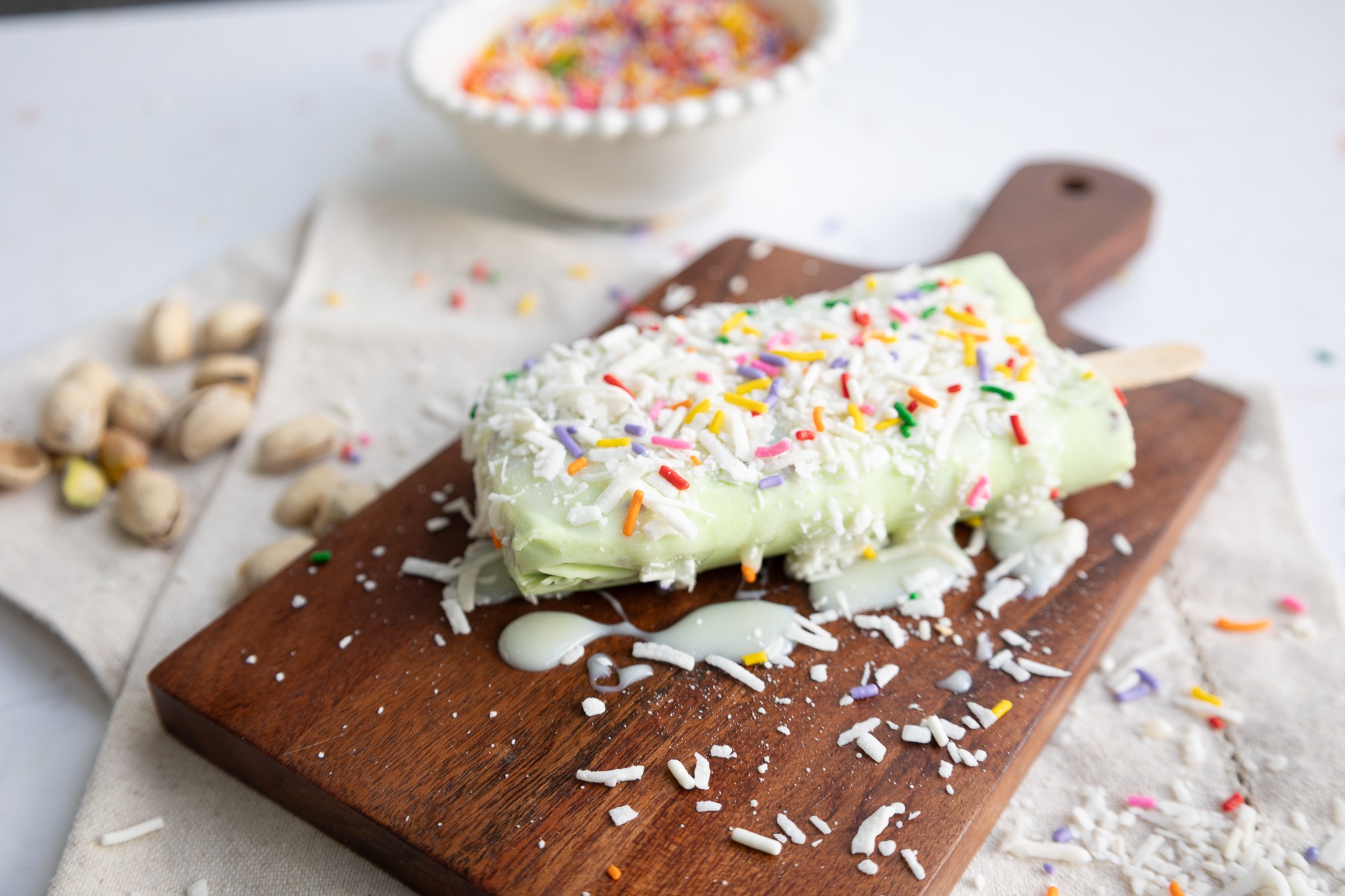 Ice cream bar with sprinkles on a wooden cutting board, surrounded by scattered sprinkles and nuts.