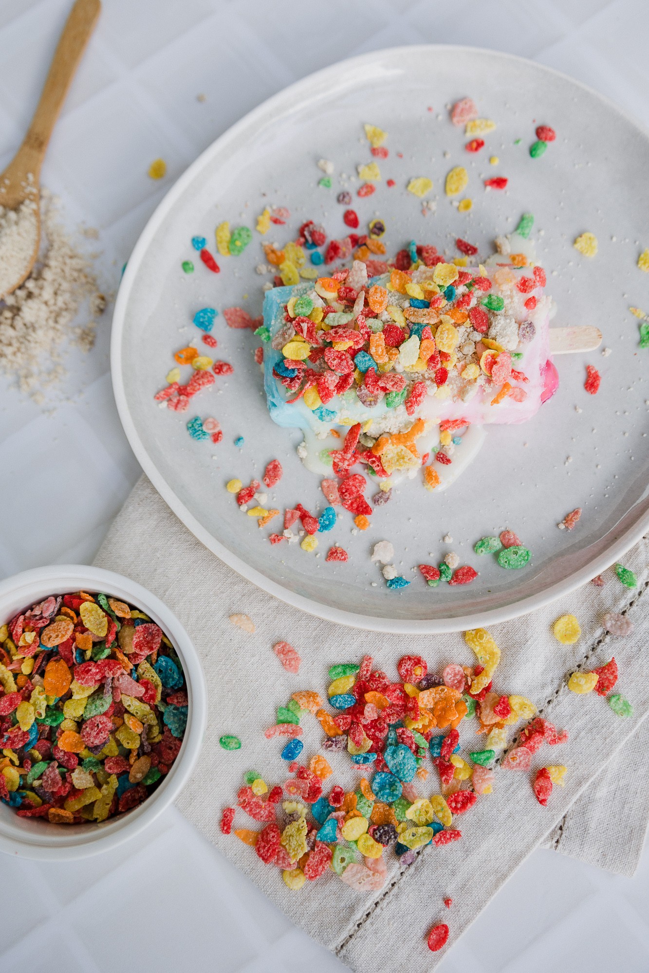 A plate with a cake topped with colorful cereal pieces, with cereal scattered on a white surface and a bowl of cereal nearby.