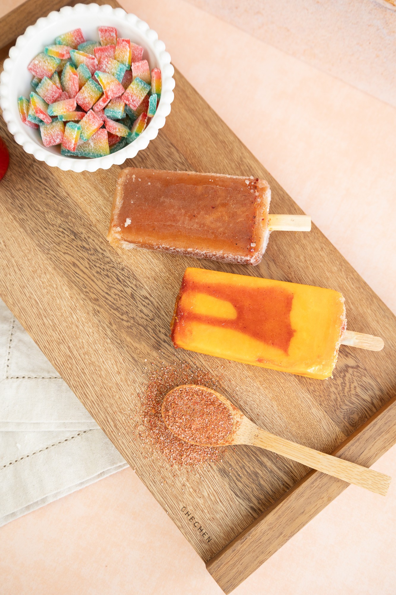 Wooden tray with colorful candies, two popsicles, and a cinnamon stick on a light surface.