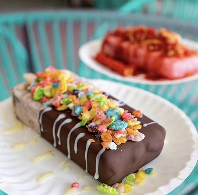 Chocolate-covered rectangular cake with colorful cereal topping and white drizzle, on a white plate.