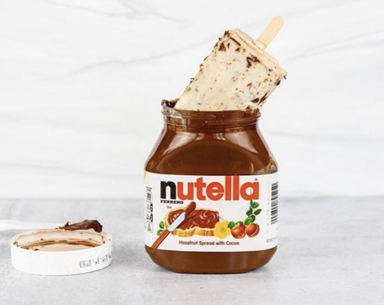 Jar of Nutella with a spoonful of spread on a white surface, lid nearby, white background.