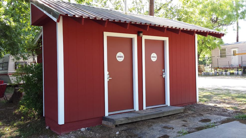 Small red building with two white doors and a sloped roof, surrounded by trees and outdoor area.