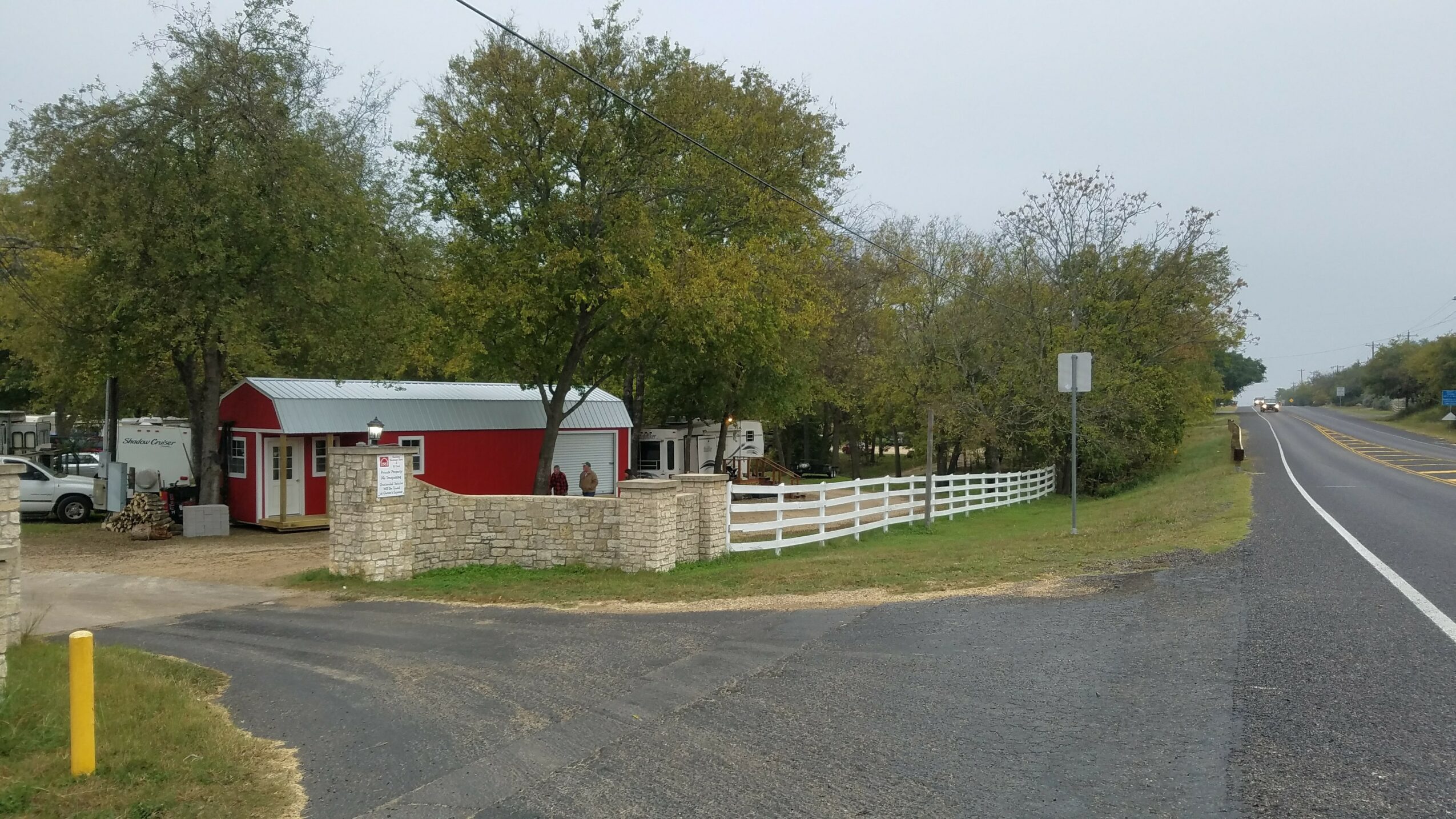 Roadside scene with trees, a small building, and a white fence, under a cloudy sky.