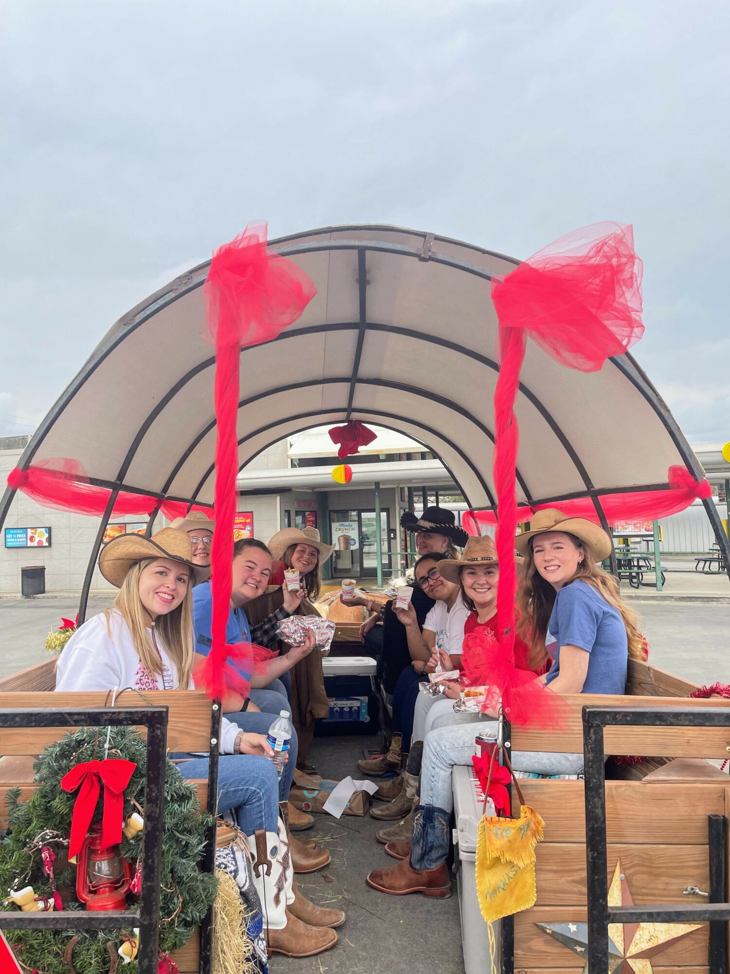 Group of people sitting inside a covered wagon, wearing cowboy hats, smiling, with decorations including red tulle and a small Christmas tree.