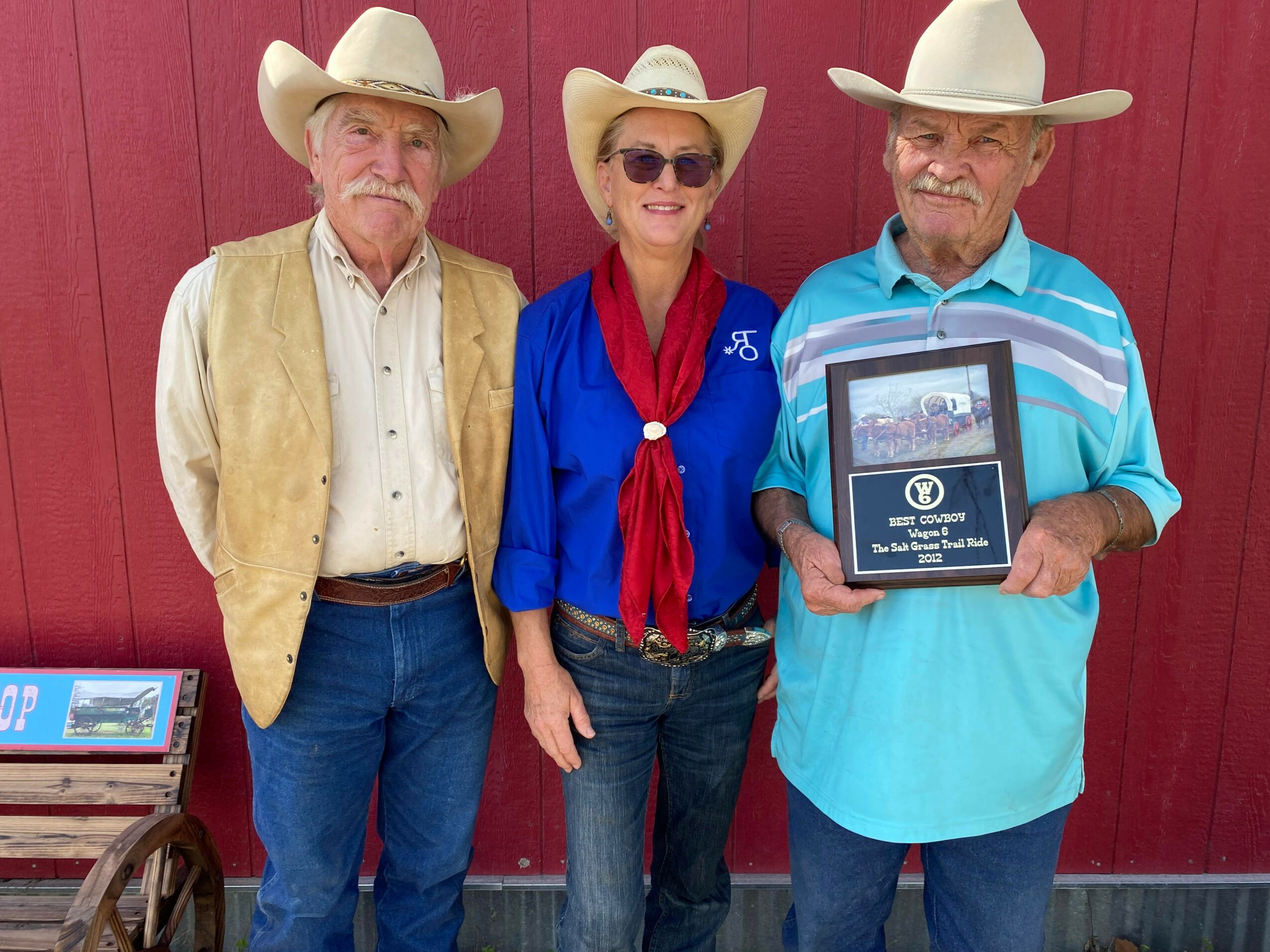 Three people wearing cowboy hats standing in front of a red wall, one holding a plaque.