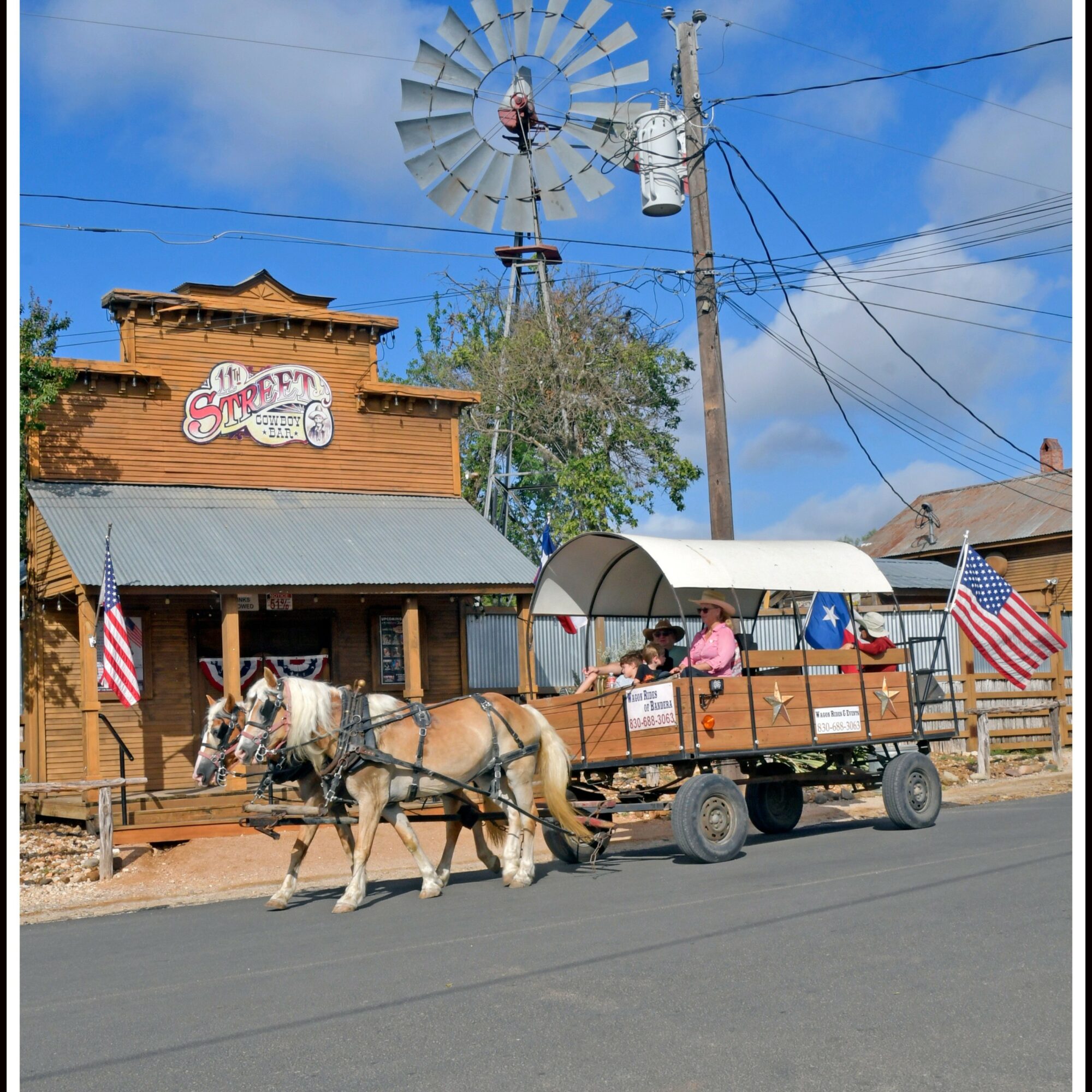 Horse-drawn wagon with people, American flags, in front of a wooden building with a sign, windmill, and blue sky.