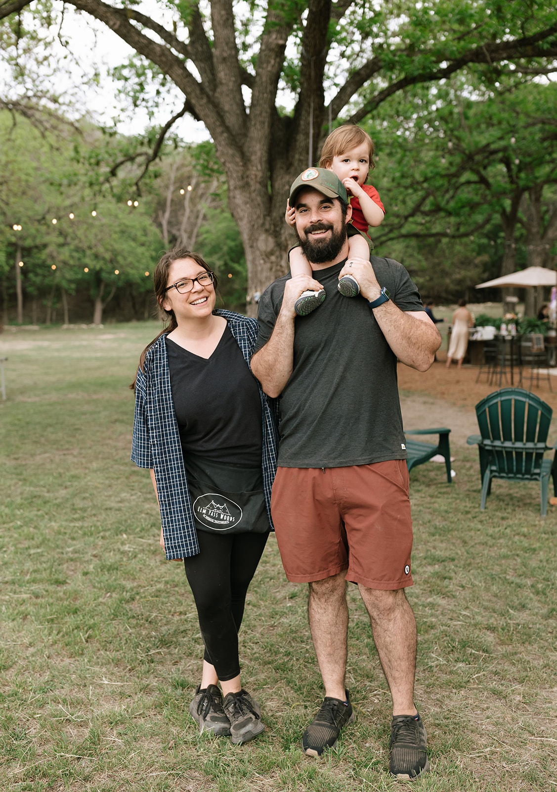 Family outdoors near a large tree, man carrying a child on shoulders, woman standing beside him, park setting.