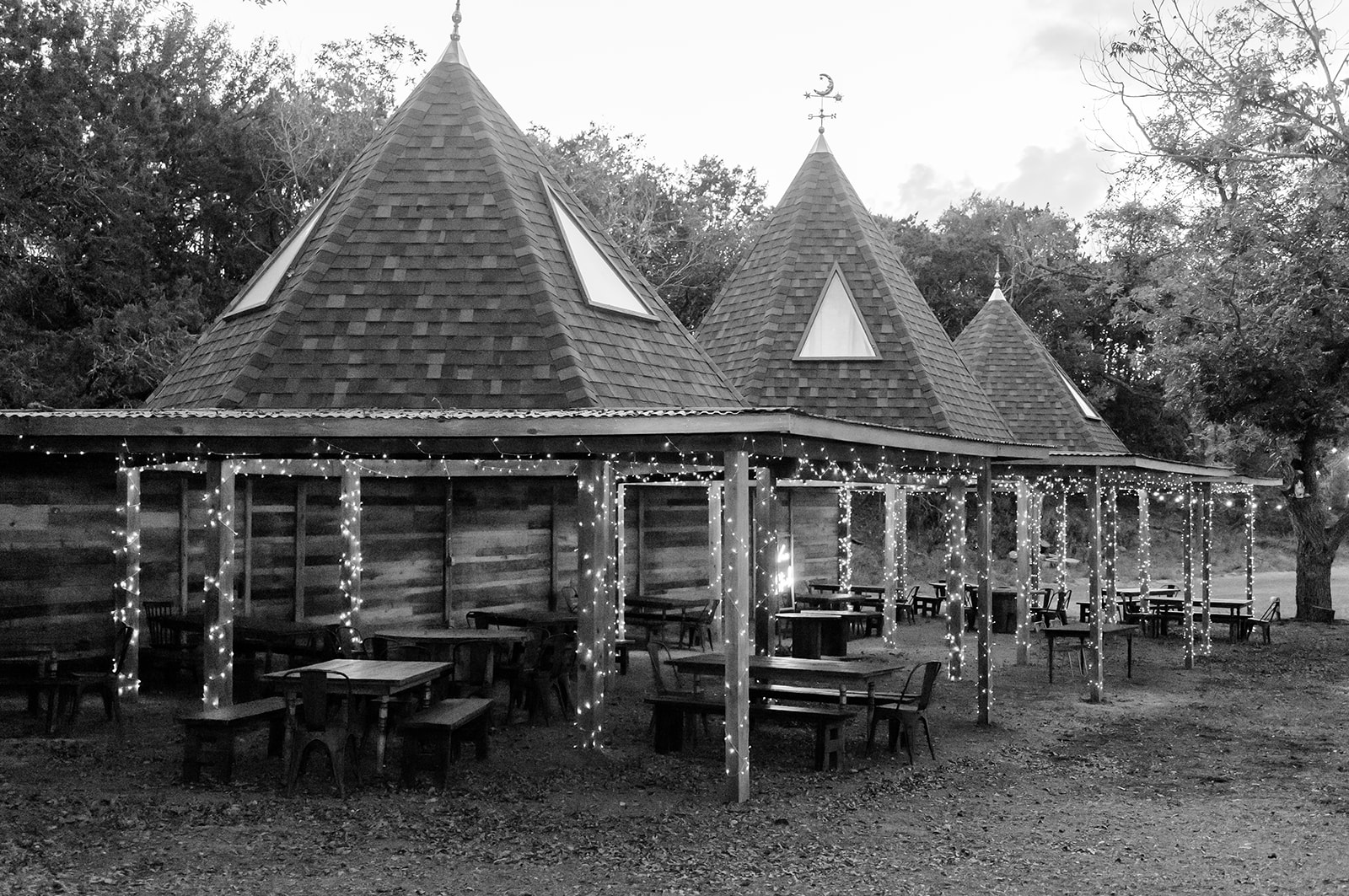 Wooden building with triangular roofs and outdoor seating area with tables and chairs, surrounded by trees.
