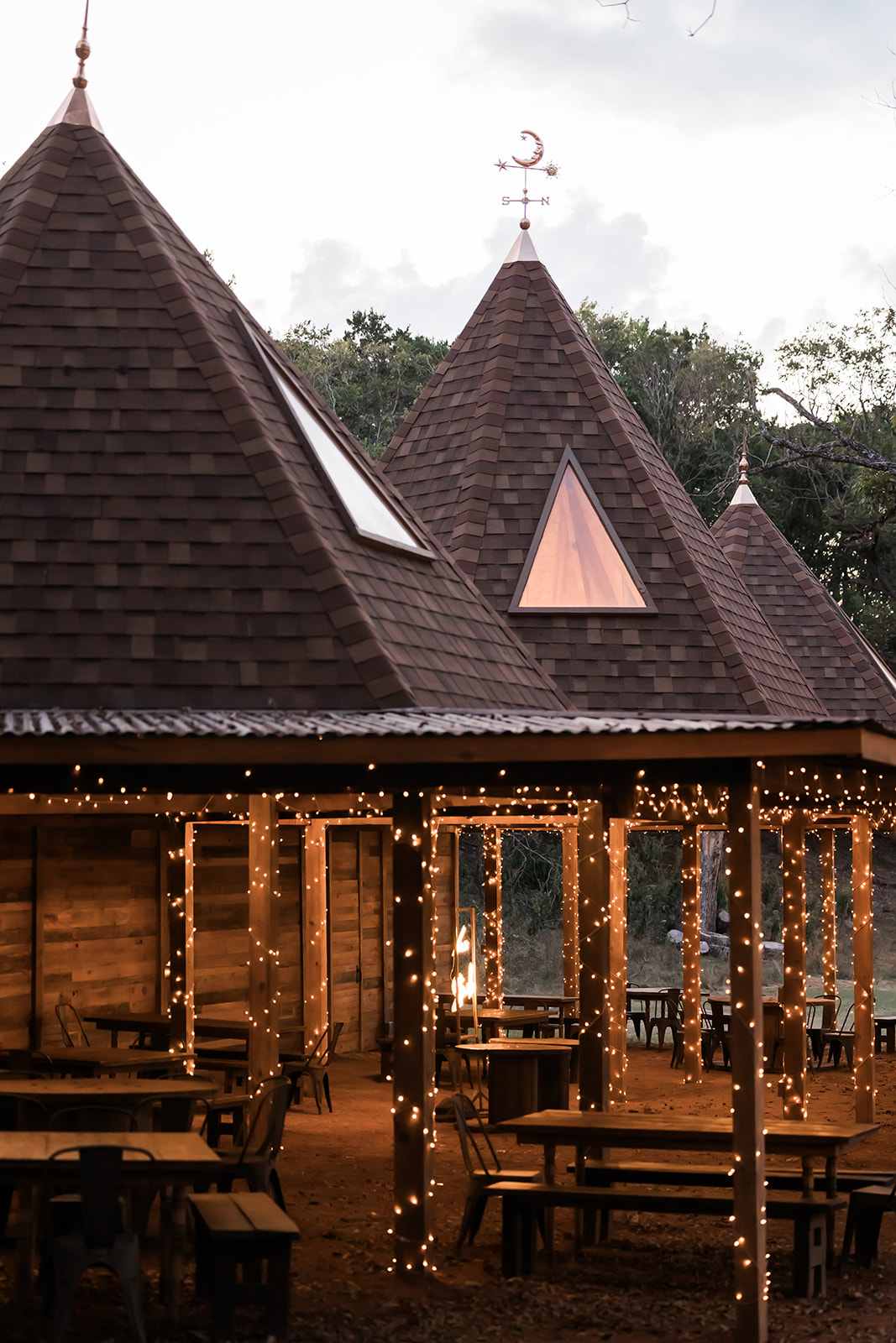 Wooden outdoor seating area with string lights in front of a building with pointed roofs and a cross on top.