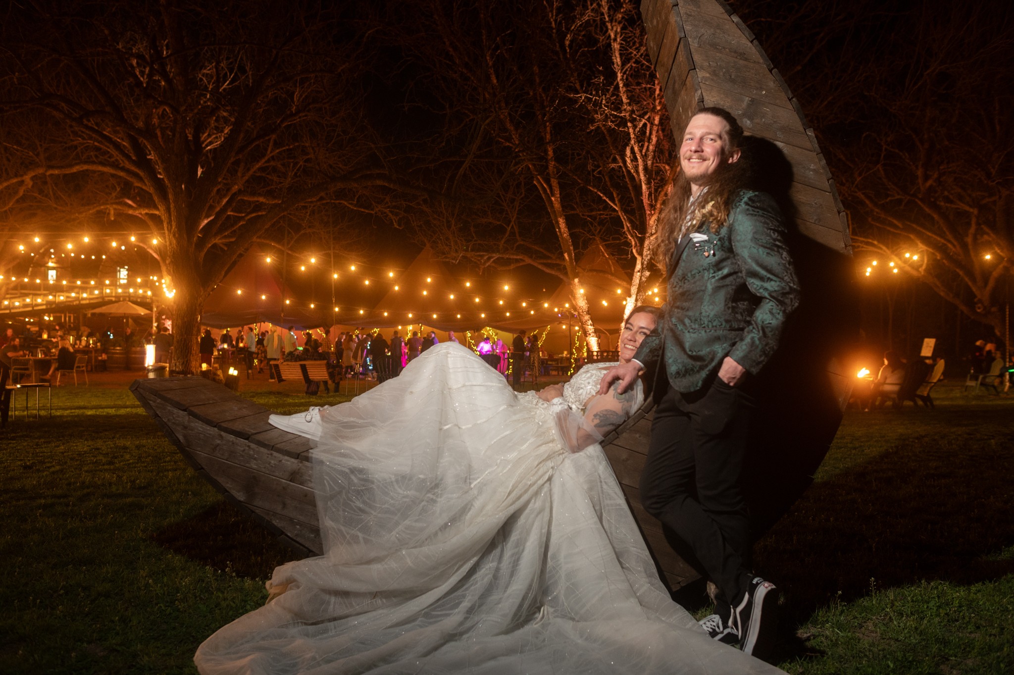 A woman in a wedding dress and a man in a jacket stand outdoors at night near a large sculpture, with string lights in the background.