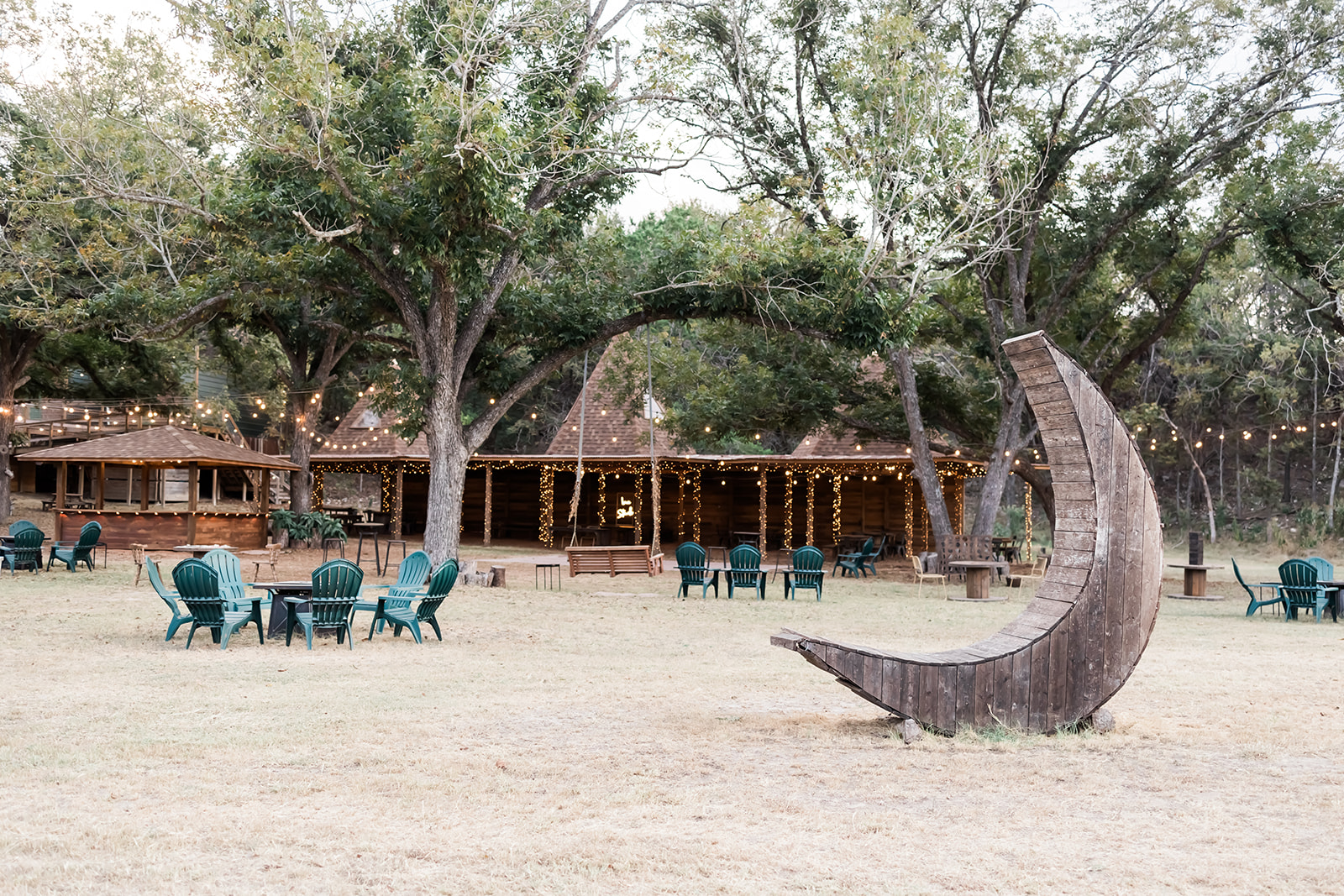 Outdoor area with trees, wooden building, and chairs, featuring a large crescent-shaped wooden sculpture in the foreground.