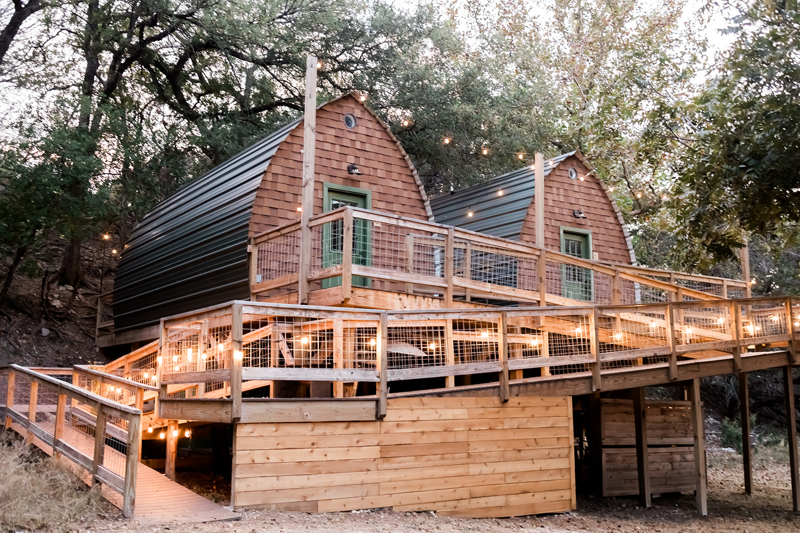 Two small, curved cabins with wooden decks and railings, surrounded by trees, illuminated by string lights at dusk.