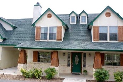 Front view of a house with green roof, dormer windows, and wooden shutters, surrounded by small bushes.