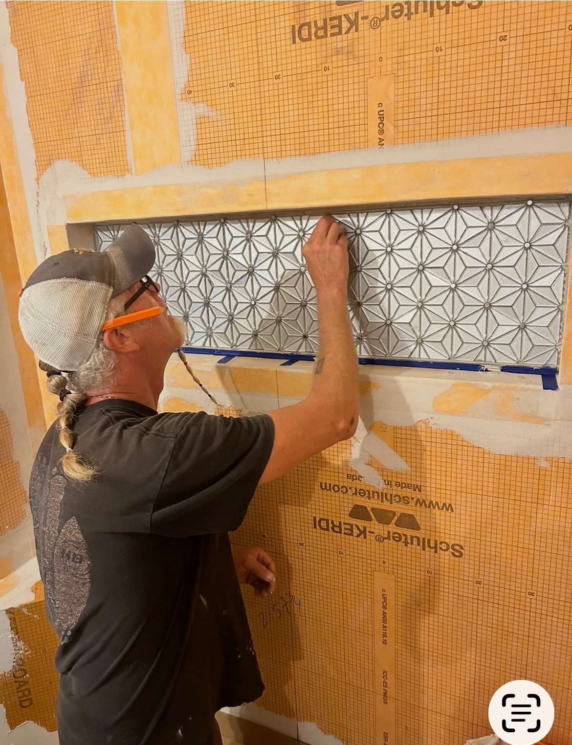 Person installing patterned tile on wall, wearing safety glasses and a cap, working on a construction site.