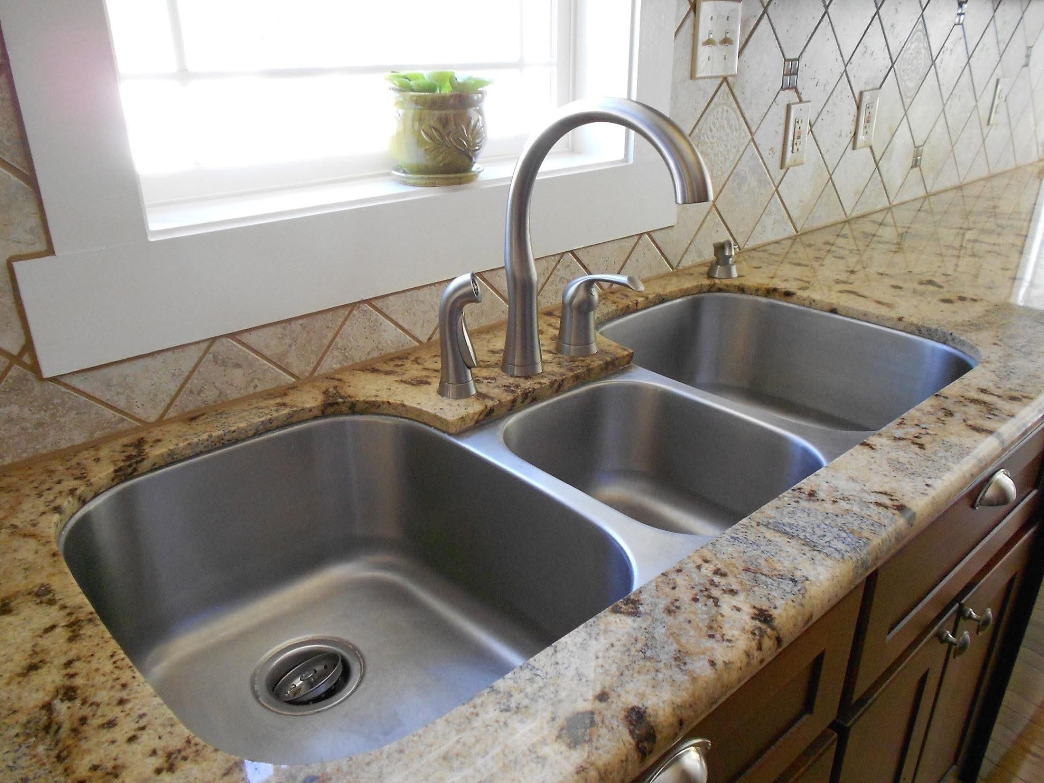 Kitchen sink with three basins, faucet, granite countertop, window, and tiled backsplash.