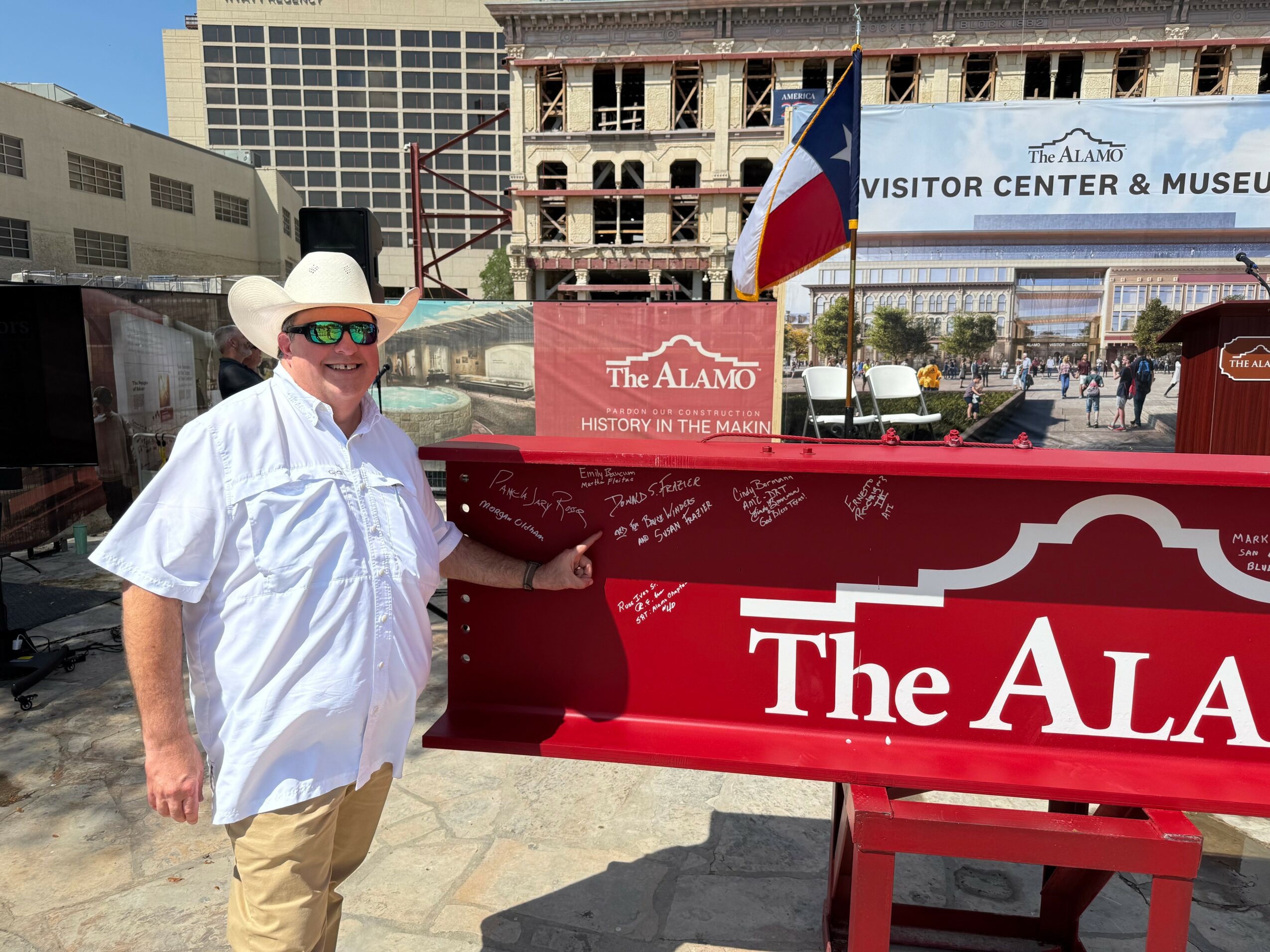 Person wearing a cowboy hat and sunglasses standing next to a red sign at an outdoor location.