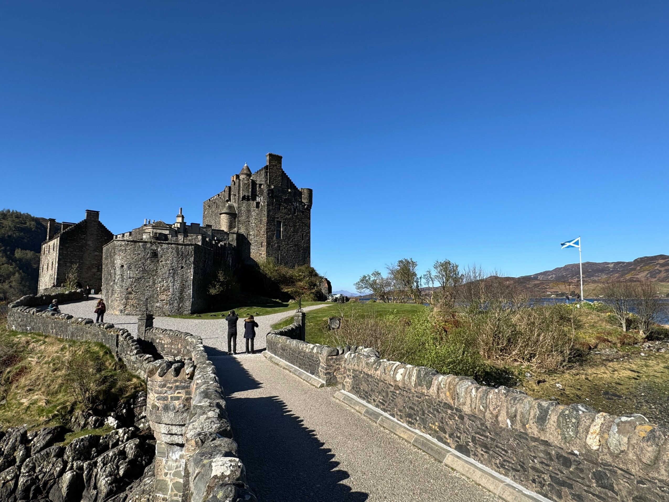 Castle on hill with stone walls and towers, pathway with people, blue sky, and landscape with trees and hills.