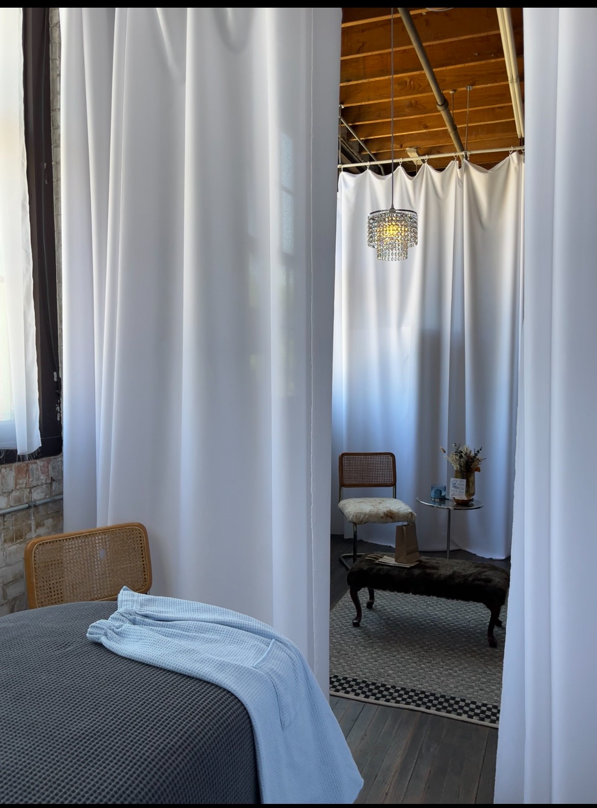 Room with white curtains, a bed with a blanket, a chair, a small table with a plant, and a chandelier.