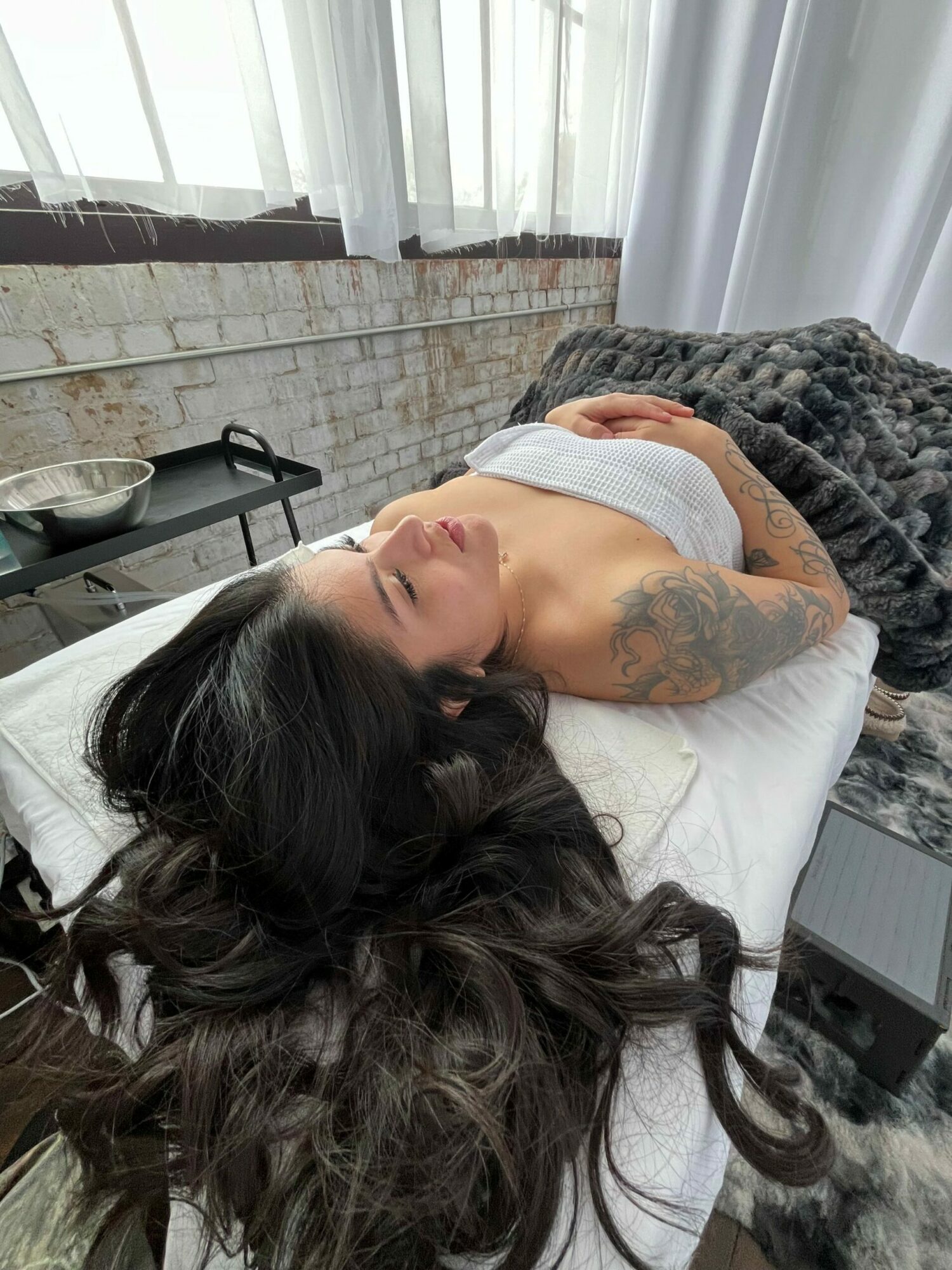 Woman with long dark hair lying on her side on a bed, eyes closed, in a bright room with white curtains and brick wall.
