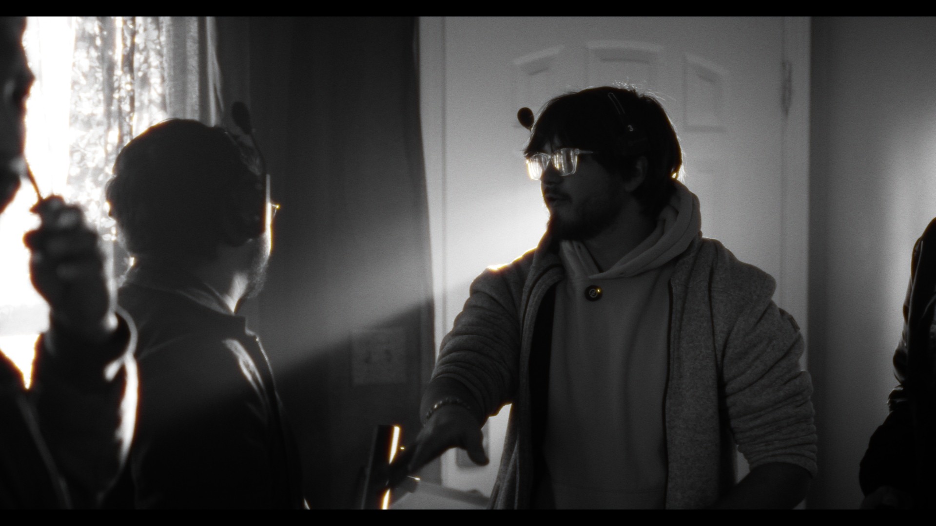 Two men face each other indoors, one with glasses and a hoodie, sunlight streaming through a window behind them.