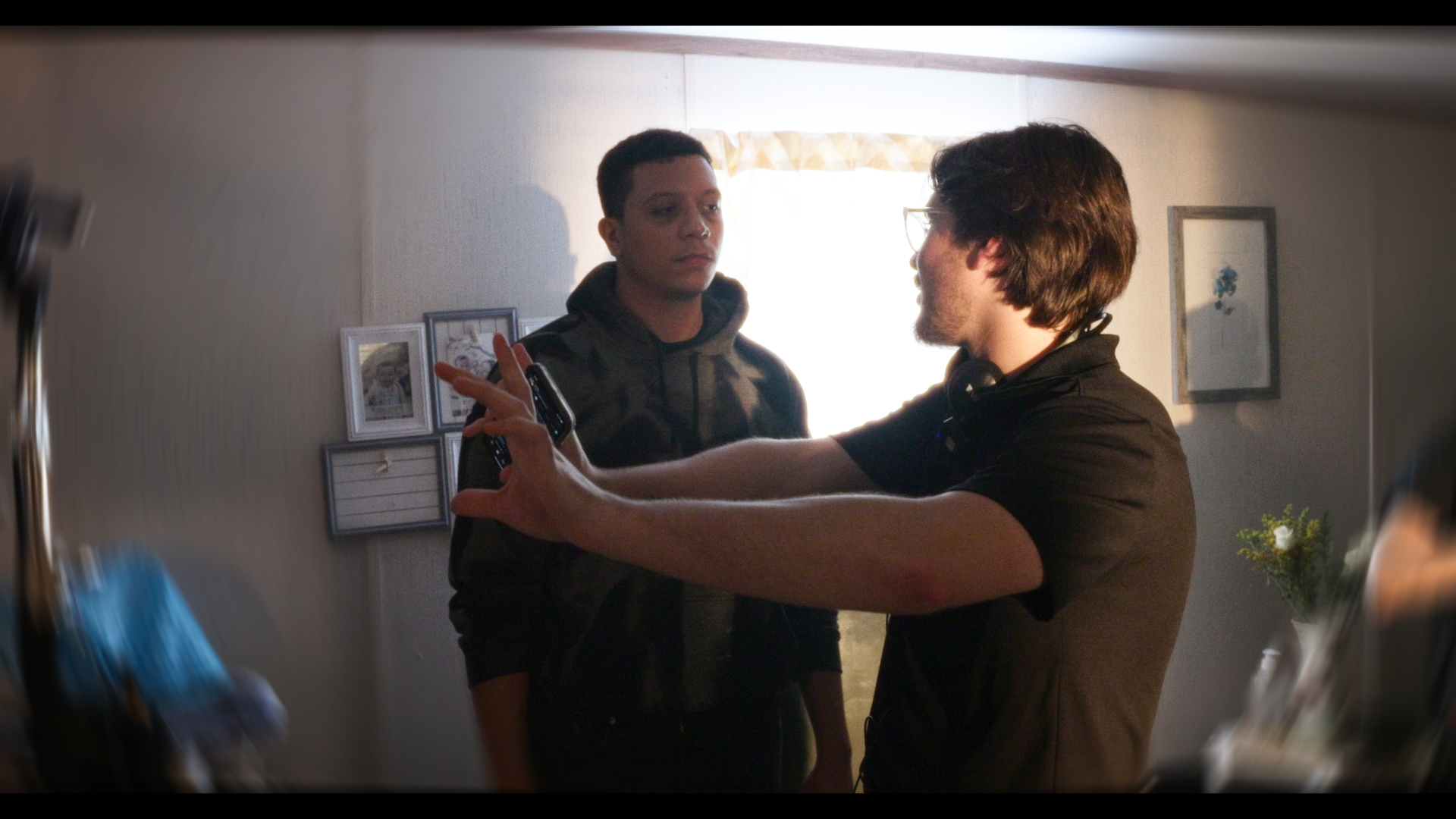 Two young men are indoors, one gesturing with hands while the other listens attentively.