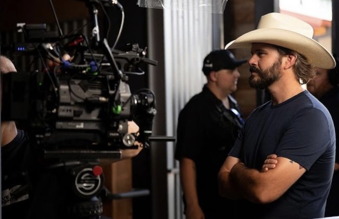 Man wearing cowboy hat and dark shirt stands with arms crossed, looking at camera equipment, in indoor setting.