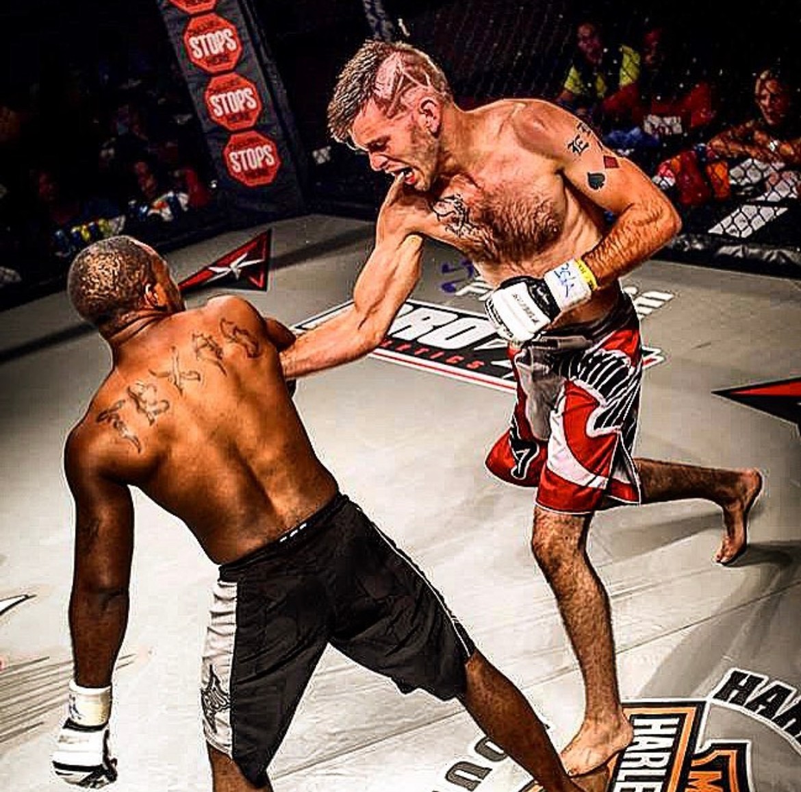 Two mixed martial artists fighting in a cage, one delivering a punch to the other's face, audience in background.