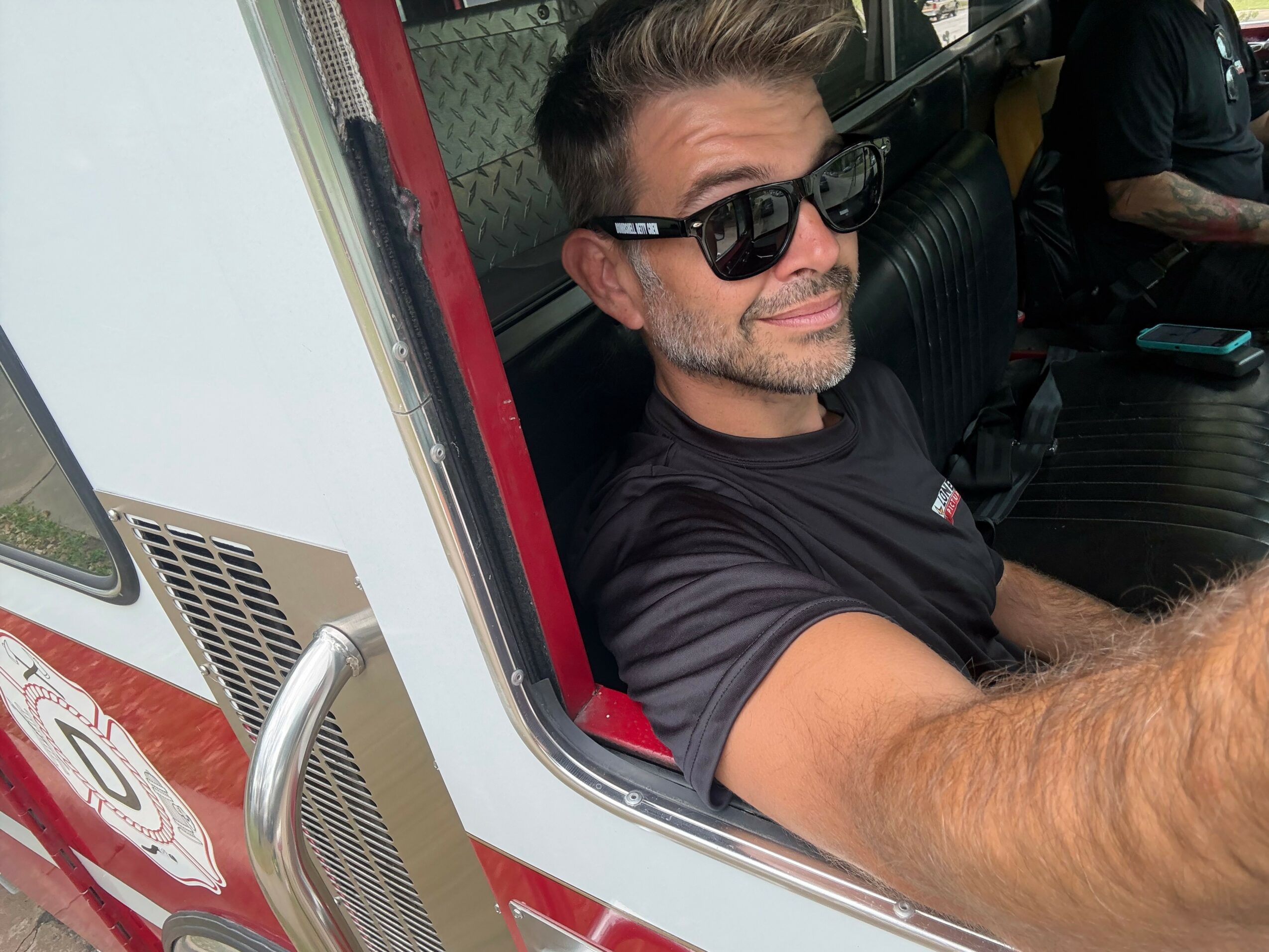 Man with sunglasses taking a selfie inside a vehicle, smiling, with a tattooed person in the background.