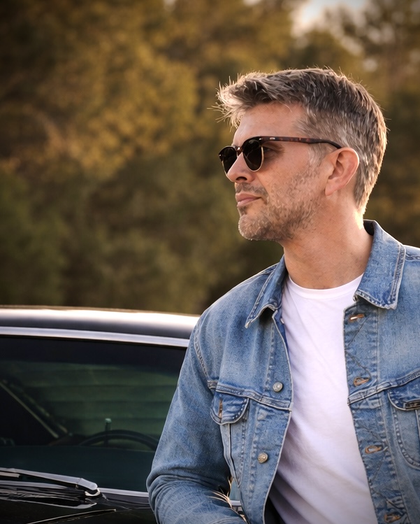 Man with sunglasses and short hair wearing a denim jacket and white t-shirt outdoors.