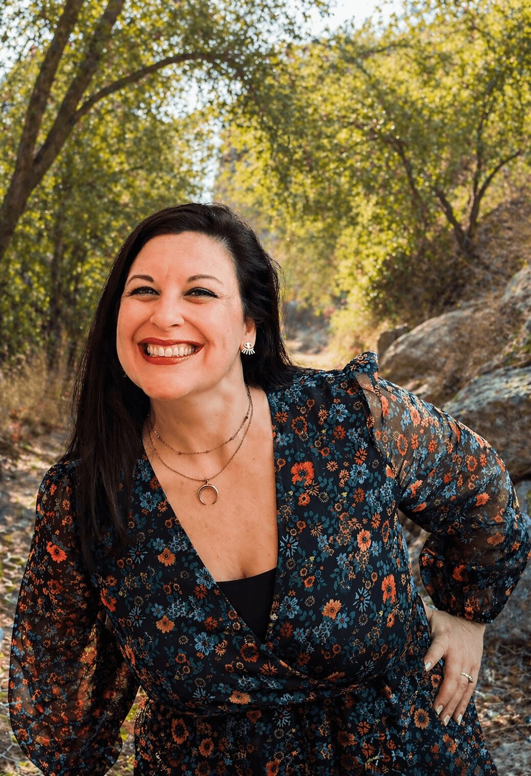 Woman with dark hair smiling outdoors, wearing a floral dress, in a natural setting with trees and rocks.