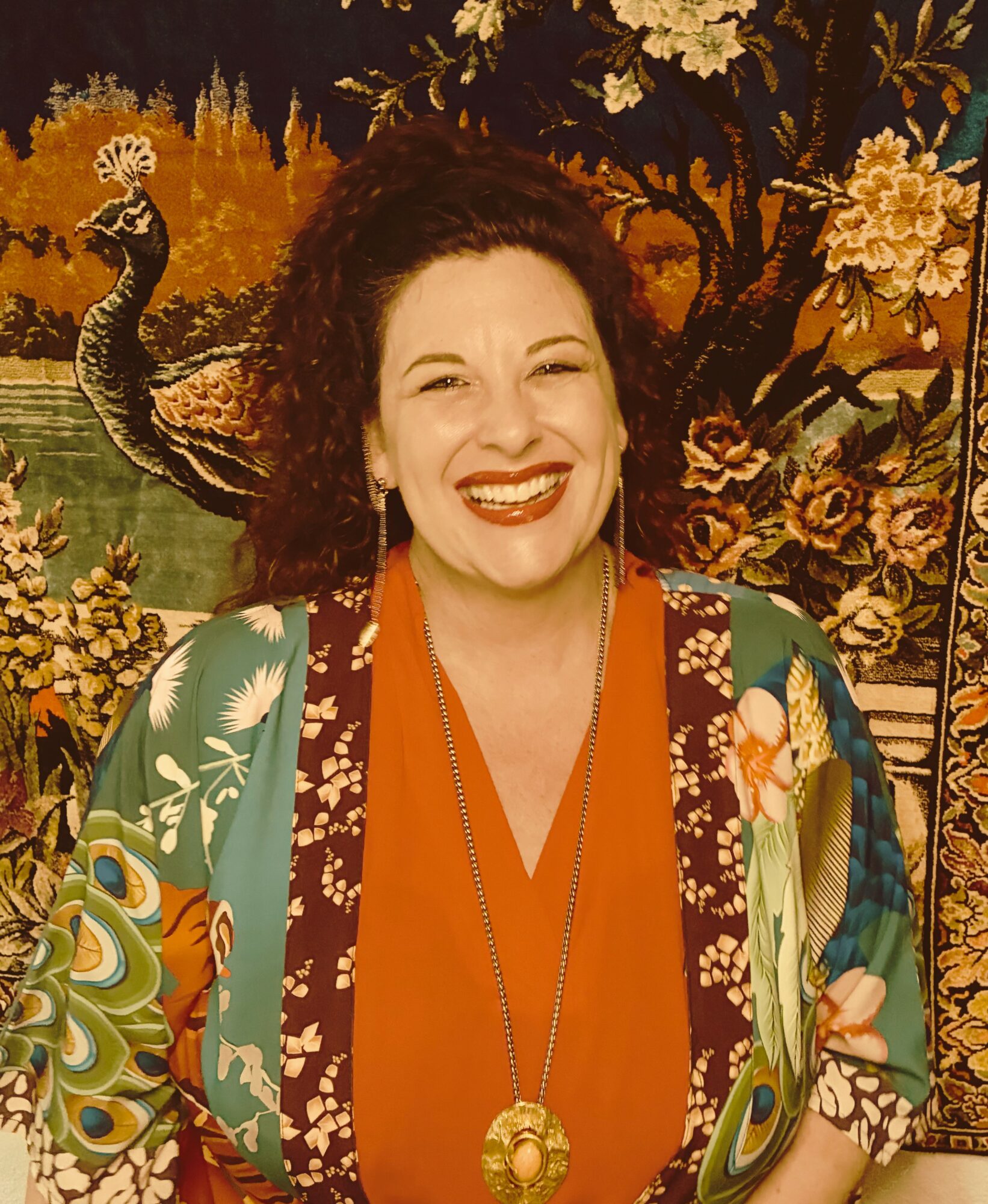 Woman with curly hair smiling, wearing an orange top and patterned jacket, in front of a colorful tapestry.