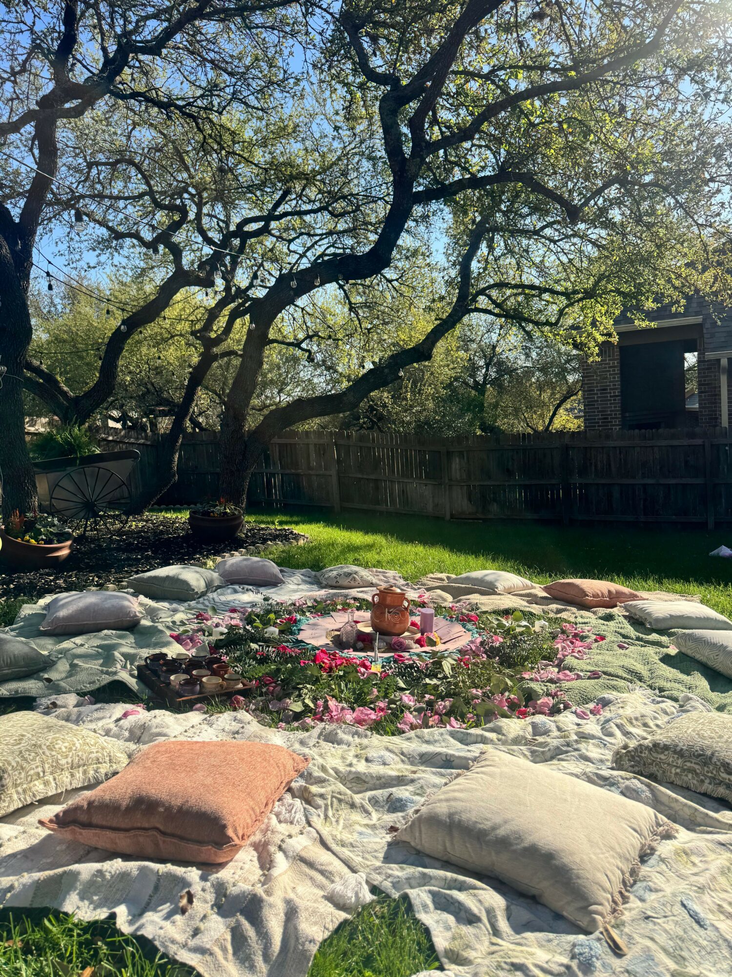 Garden with trees, flowers, rocks, and a blanket, viewed from the side, with a fence and house in the background.